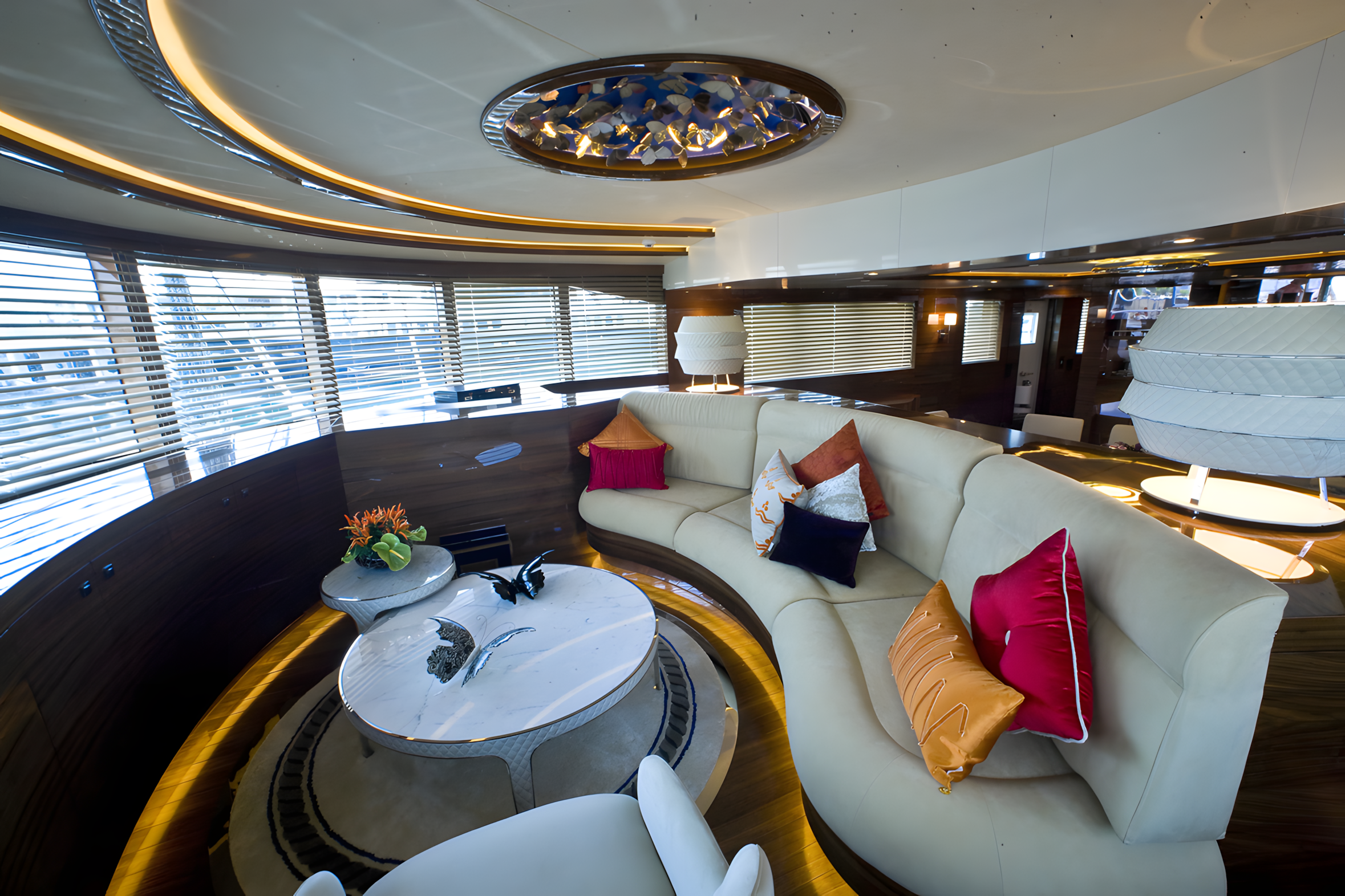 Interior of a luxurious yacht with a curved white sofa decorated with colorful pillows, a marble coffee table with a butterfly sculpture, and large windows with blinds, offering a view of the city skyline.