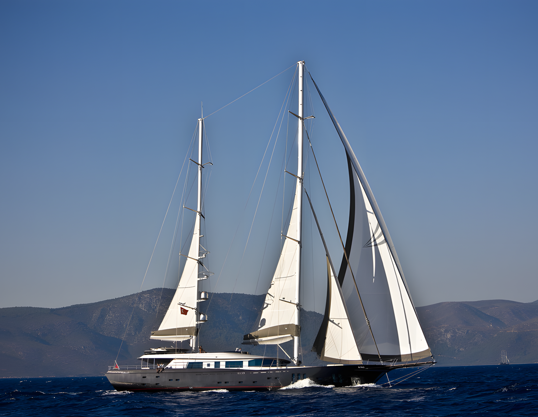 Sailing yacht with white sails on blue ocean with mountainous land in background.
