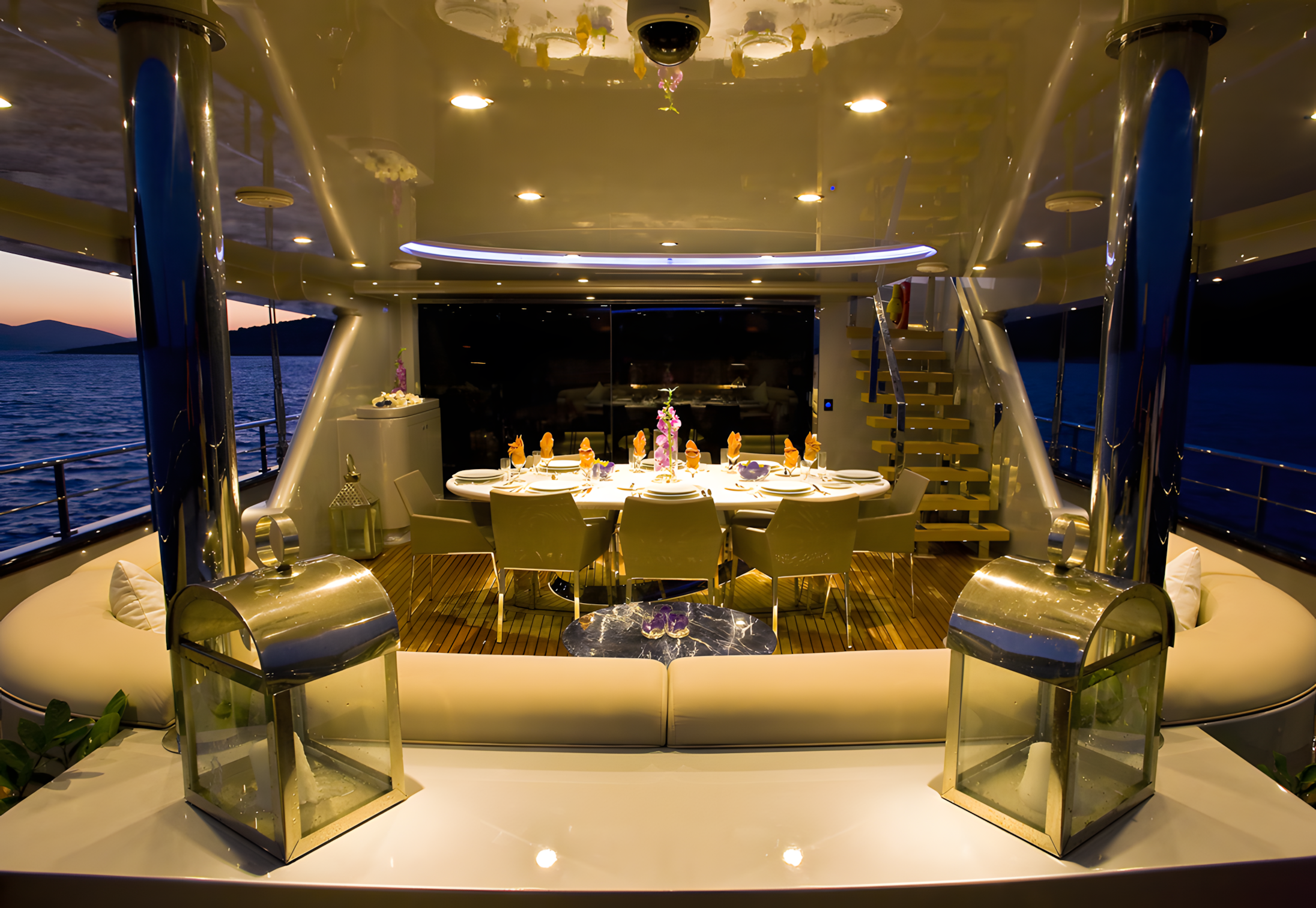 An elegantly set dining table on a yacht with a view of the ocean at sunset, decorated with flowers and candles, surrounded by modern furniture and lighting.
