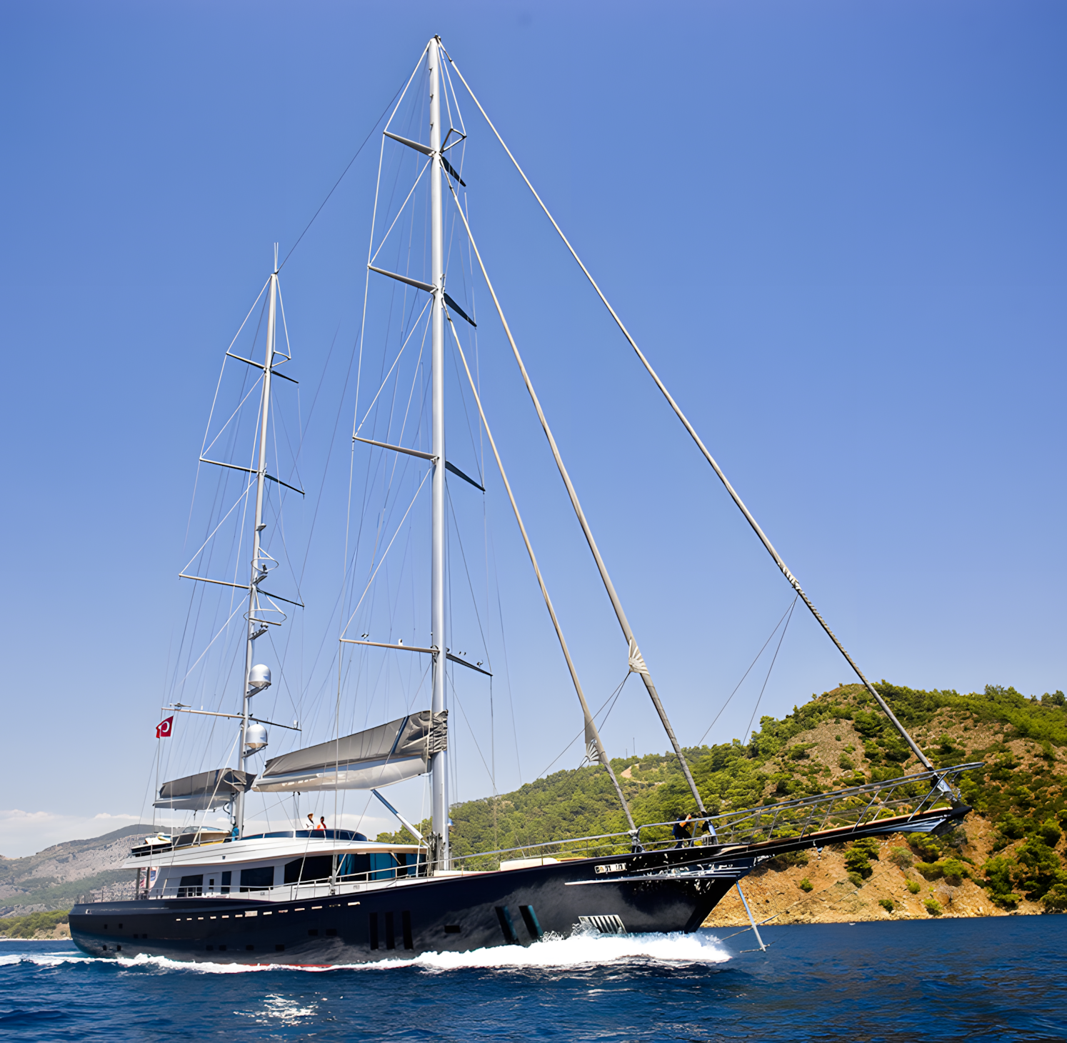 A large black sailing yacht with two tall masts sailing on a calm sea near a green hilly coastline under a clear blue sky.