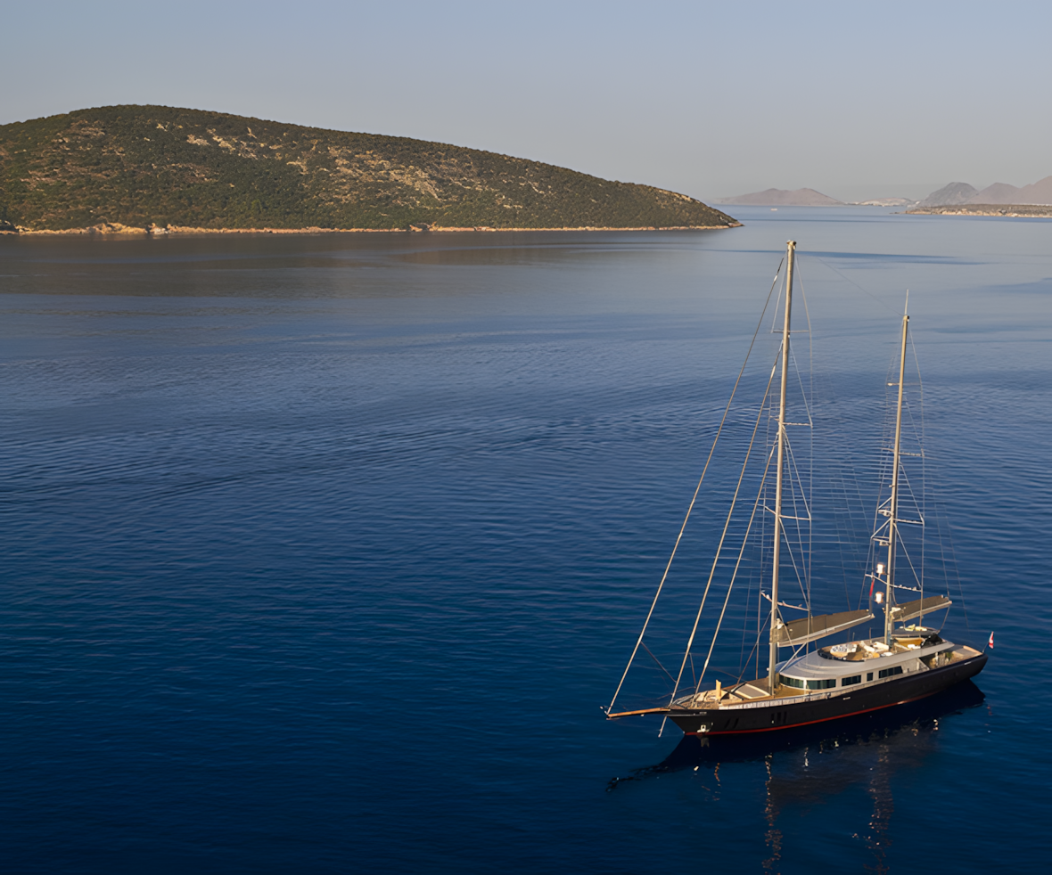 A sailboat floating on calm blue water near a green, hilly island with distant mountains in the background.