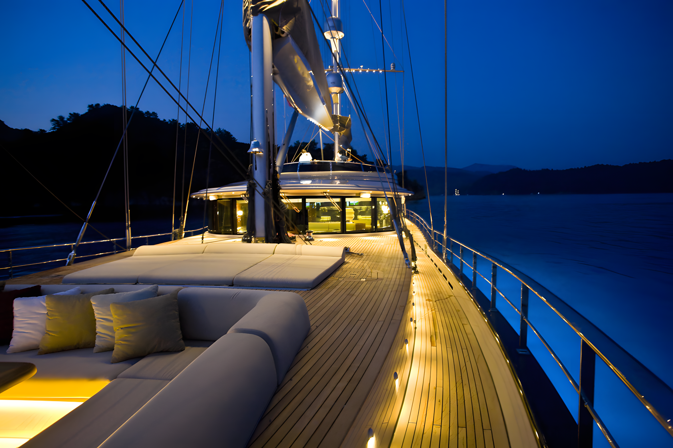 Luxury yacht deck at night with cushioned seating, string lights, and a view of water and mountains in the background.