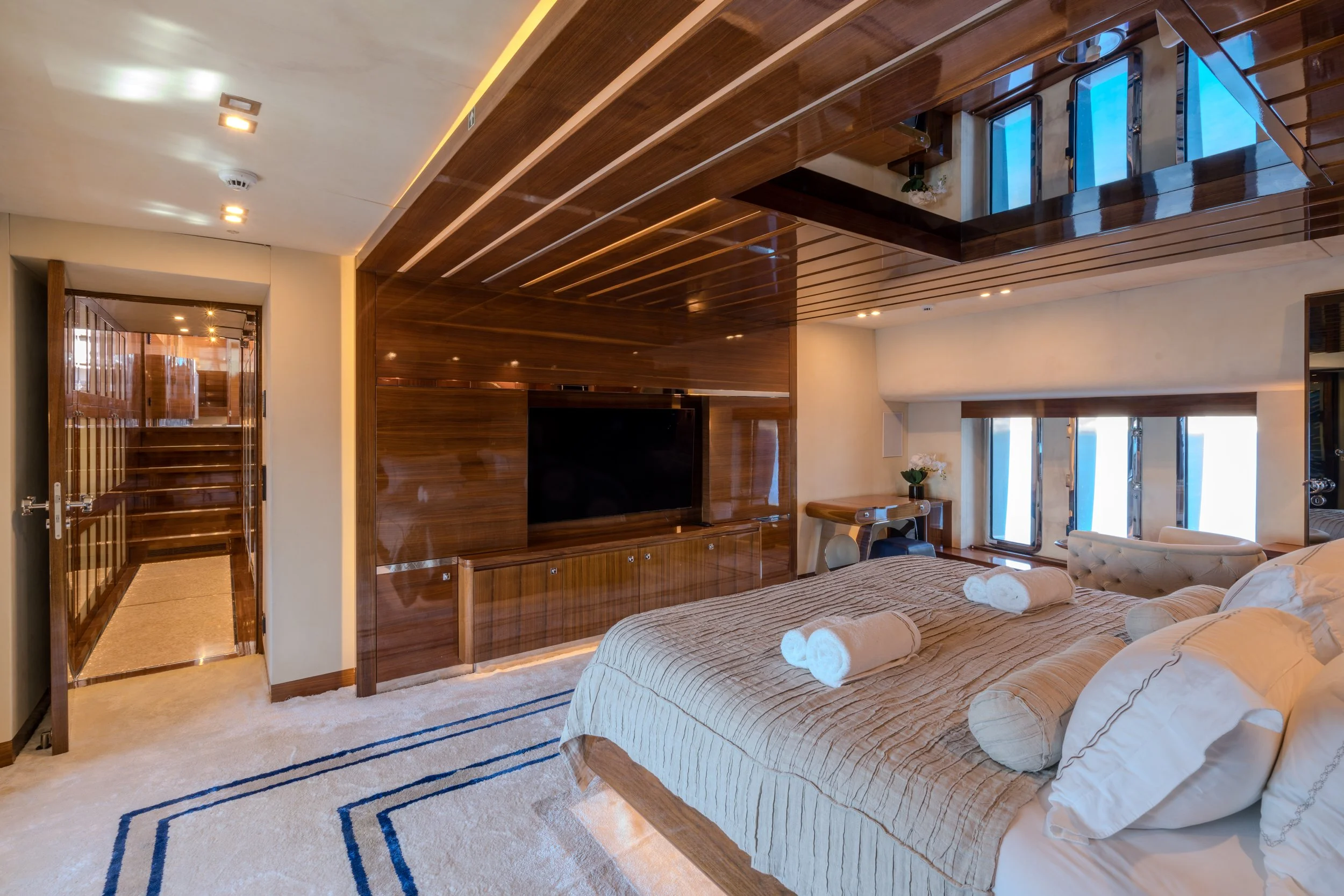 A luxurious bedroom featuring a large bed with rolled towels, a flat-screen TV on a polished wood wall, and a window with a view. The room has a cream-colored carpet, a small desk with a chair and a vase of white flowers, and a mirrored ceiling refle