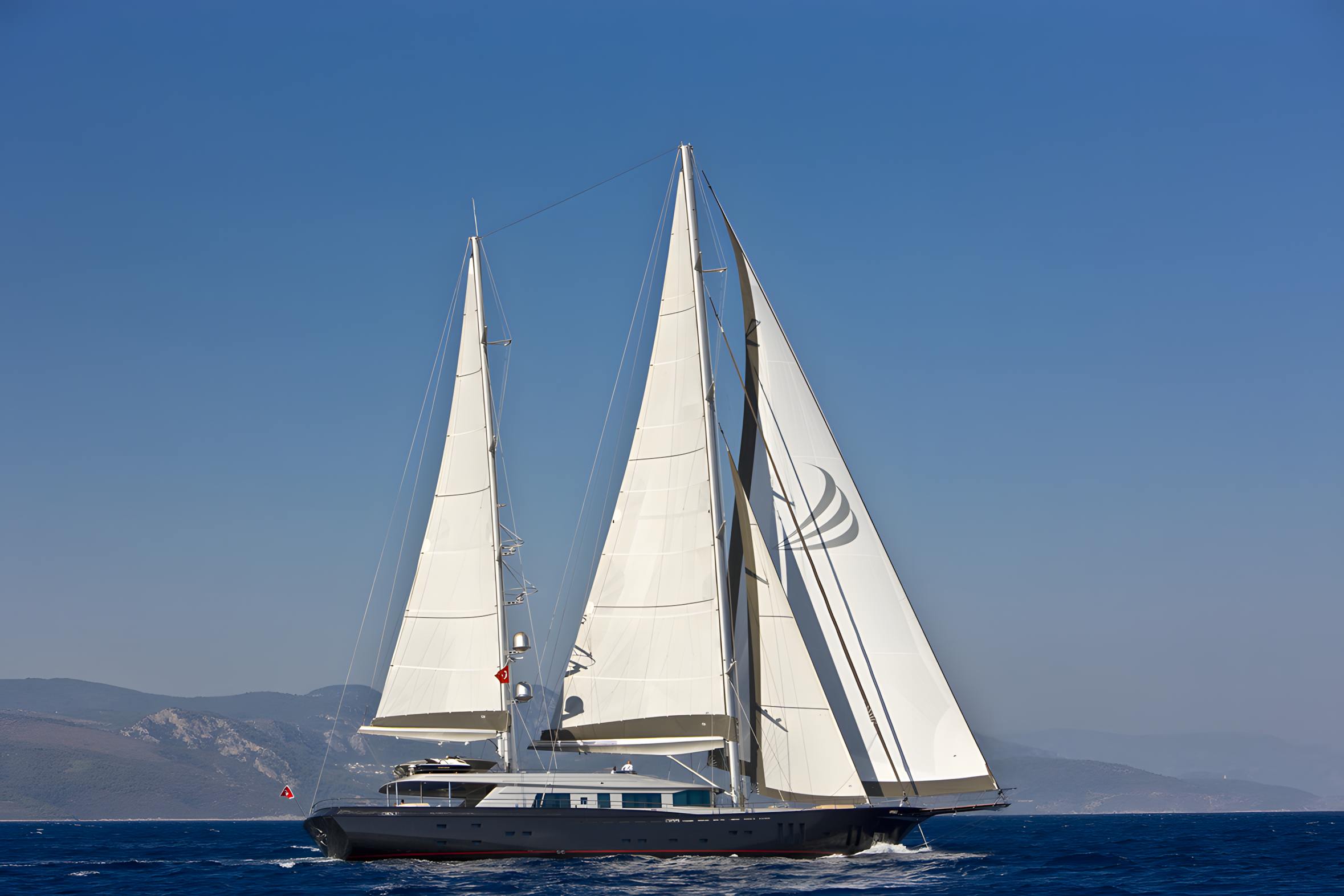 A large sailing yacht with white sails on the open ocean, mountains in the background, under a clear blue sky.