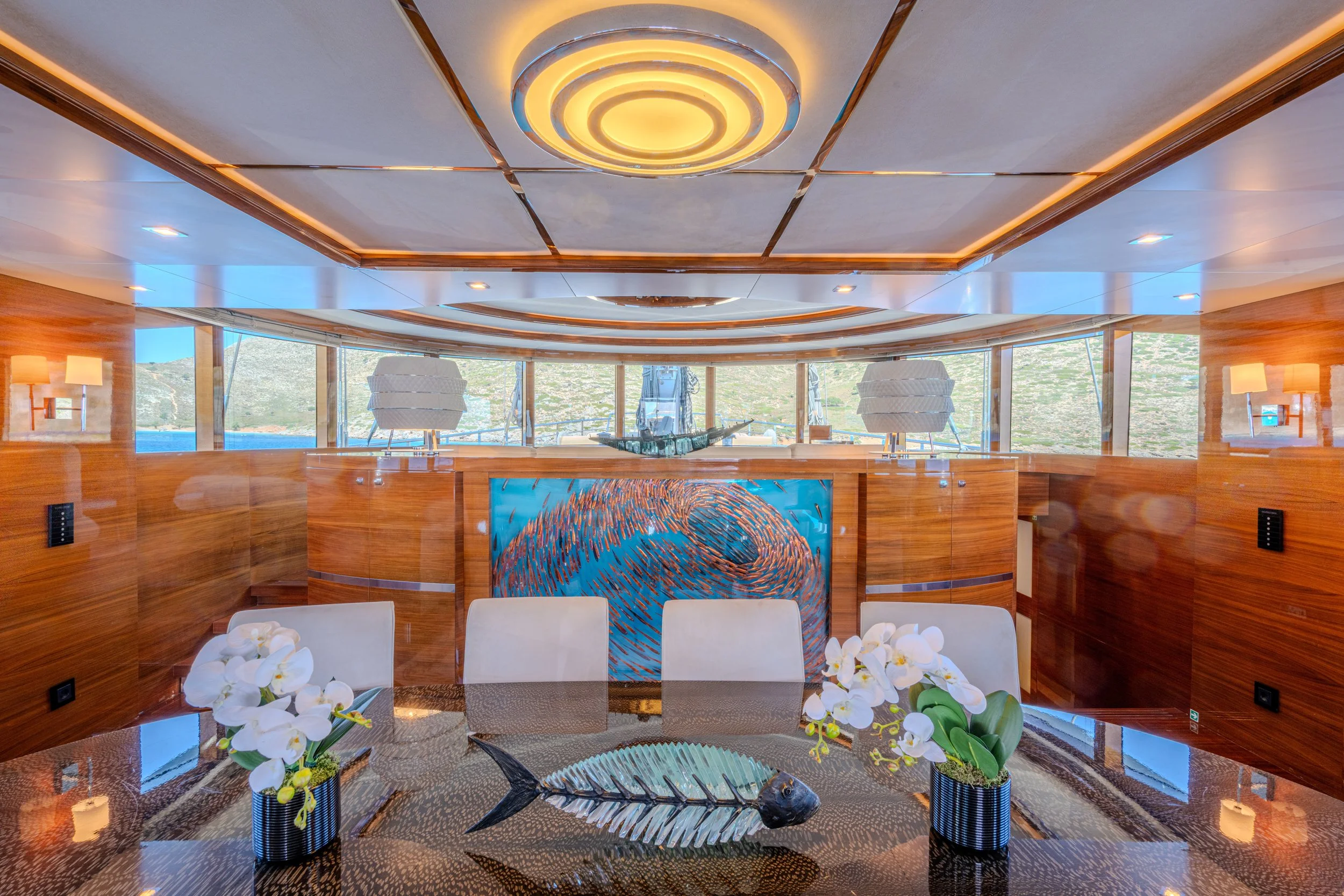 Interior of a luxurious yacht dining room with a glass table, white orchids, a fish sculpture, wood-paneled walls, and windows showing a scenic coastal view.