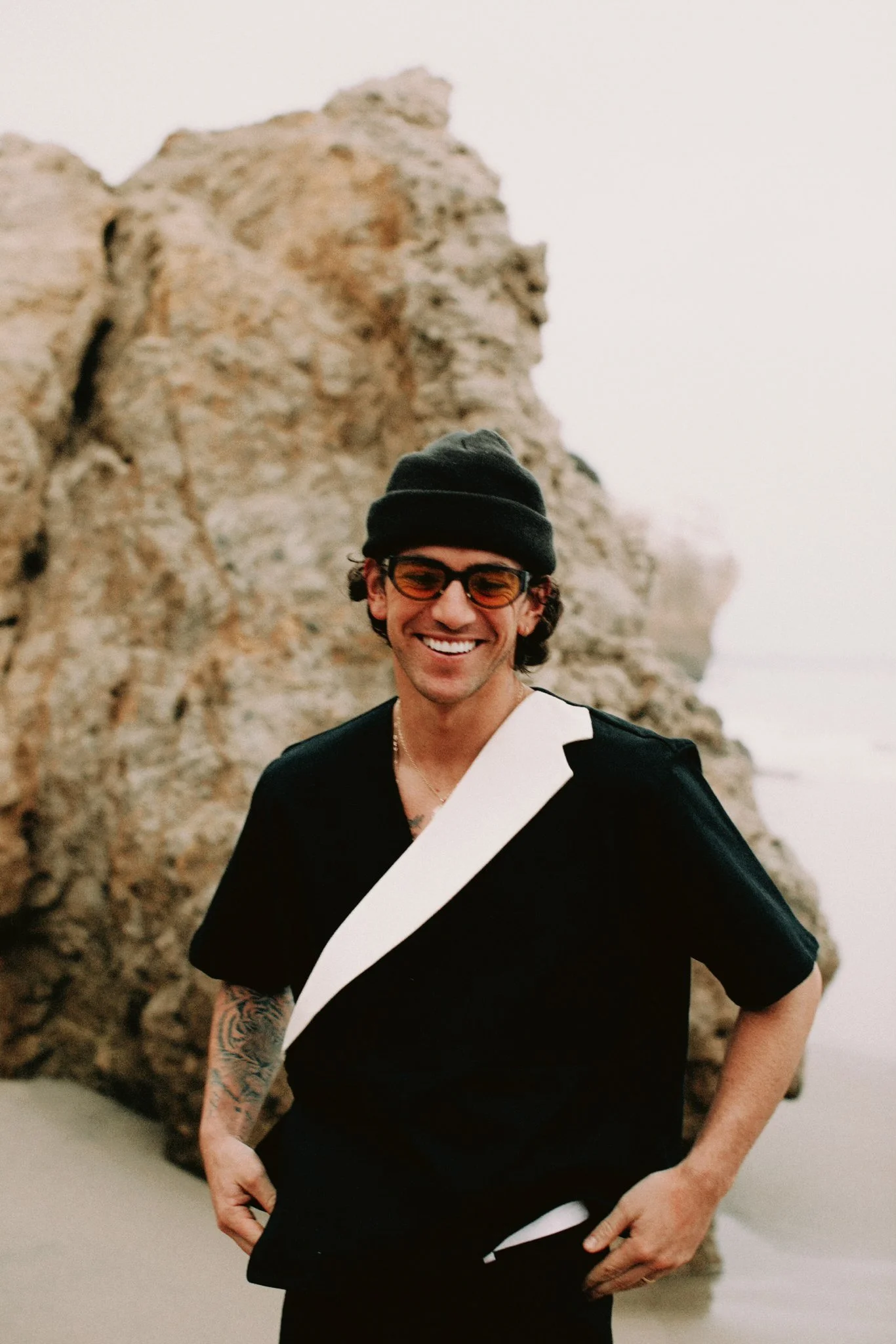Smiling man with dark hair, glasses, and a beanie, standing outdoors near rocks and foggy weather.