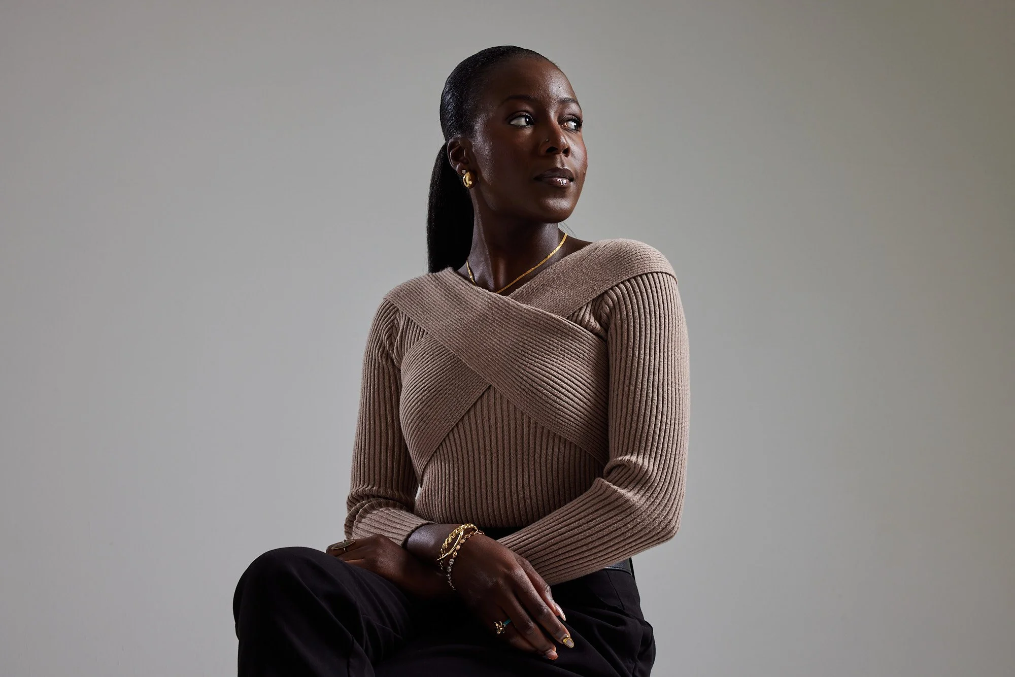 A woman with dark skin and long dark hair styled in a ponytail, wearing a beige ribbed sweater and black pants, sitting against a plain gray background, looking to the side with her arms crossed.