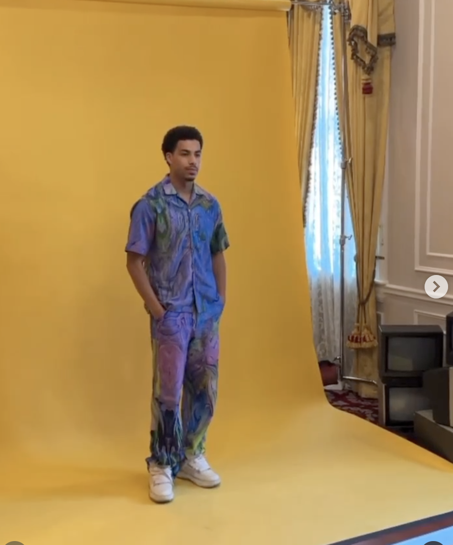 Young man standing with hands in pockets against a yellow photo backdrop in a room with gold curtains and a television.