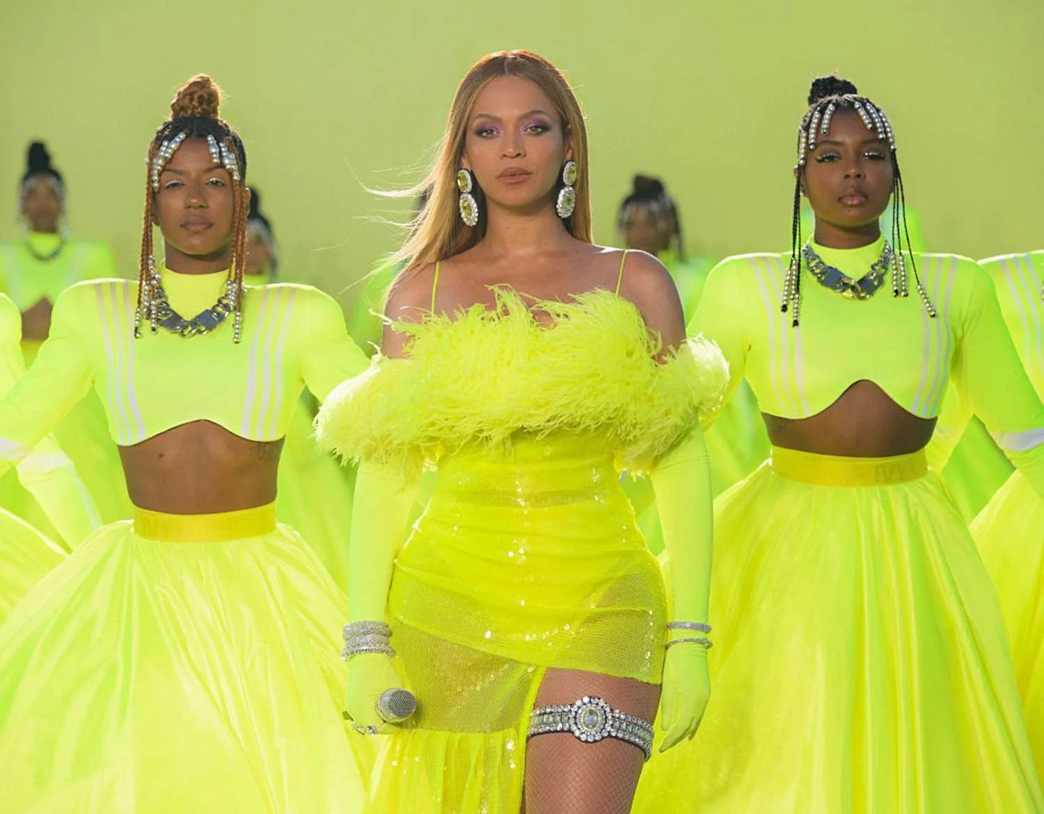 A woman in a bright yellow feathered dress stands in the center of a group of women wearing matching yellow outfits, all on a green background.