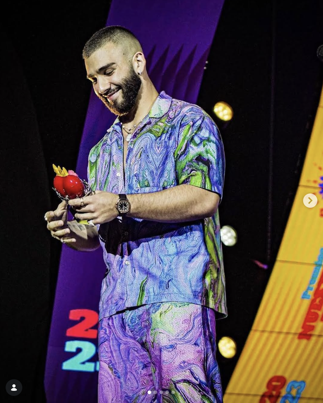 A man with a beard and short hair, wearing a colorful, abstract-patterned shirt and matching pants, is holding a red apple-shaped award with a small yellow crown on top. He is smiling and looking down at the award, standing on a stage with purple and yellow background elements.