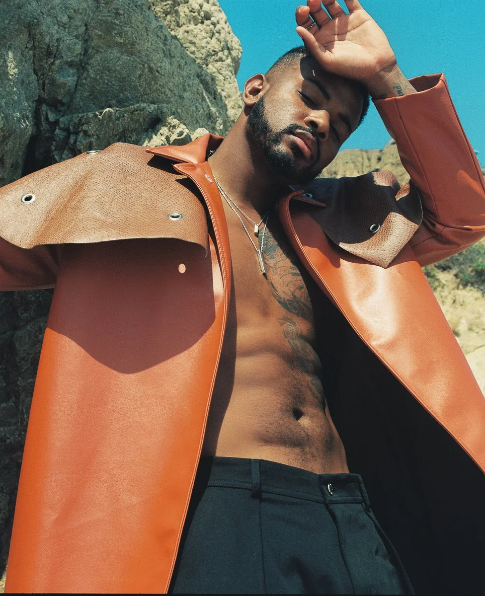 A shirtless man wearing a tan and orange jacket outdoors with rocky terrain in the background, shielding his eyes from the sun with one hand.