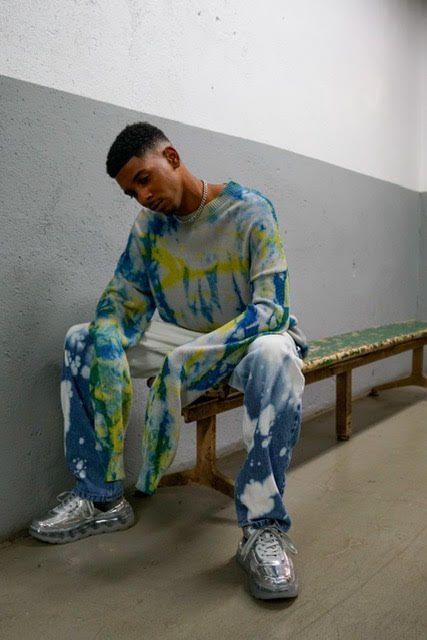 Young person sitting on a wooden bench against a gray and white wall, wearing a colorful tie-dye hoodie and matching jeans with silver sneakers.