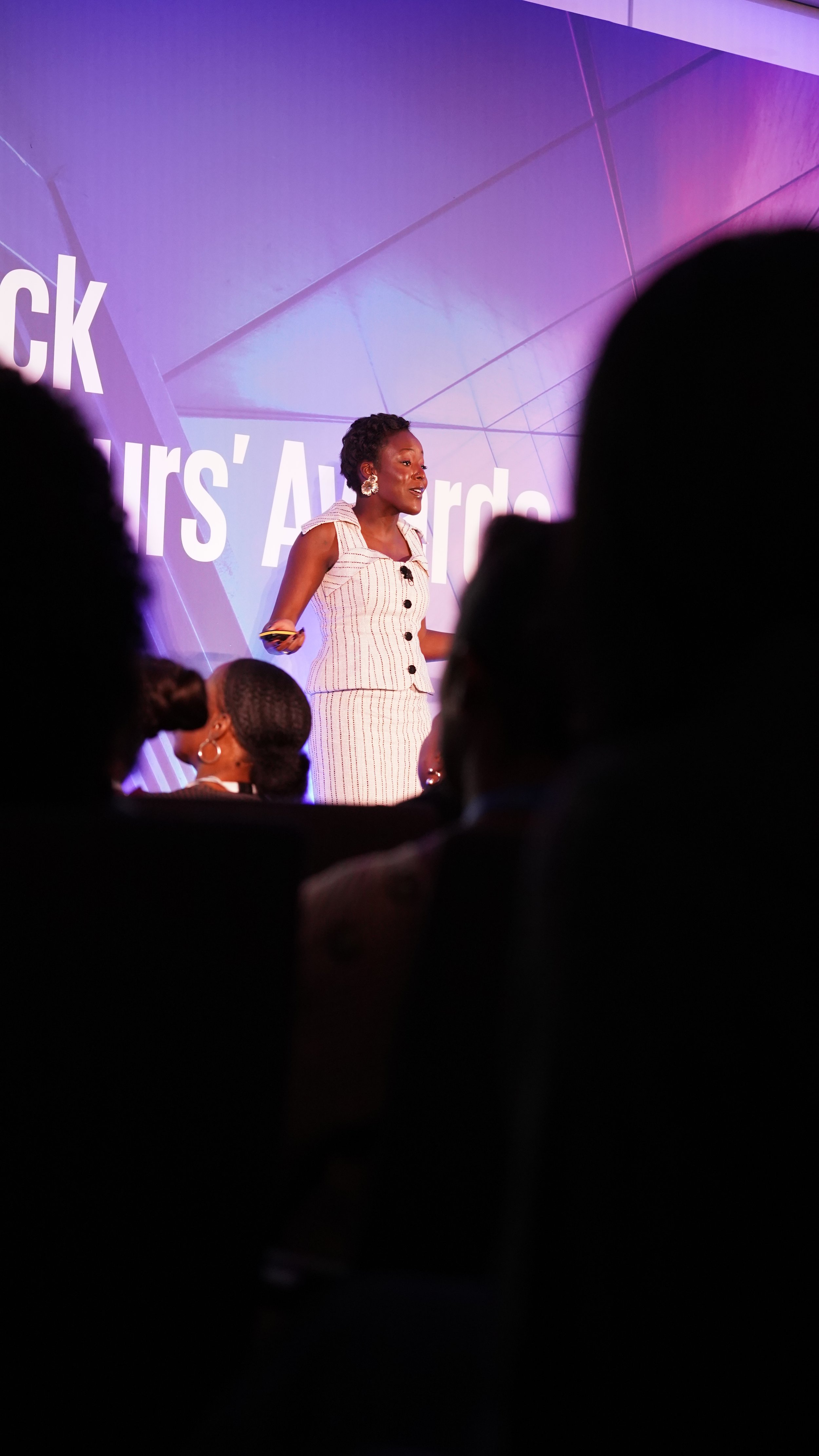 A woman speaking on stage at an awards event with a large screen behind her displaying text.