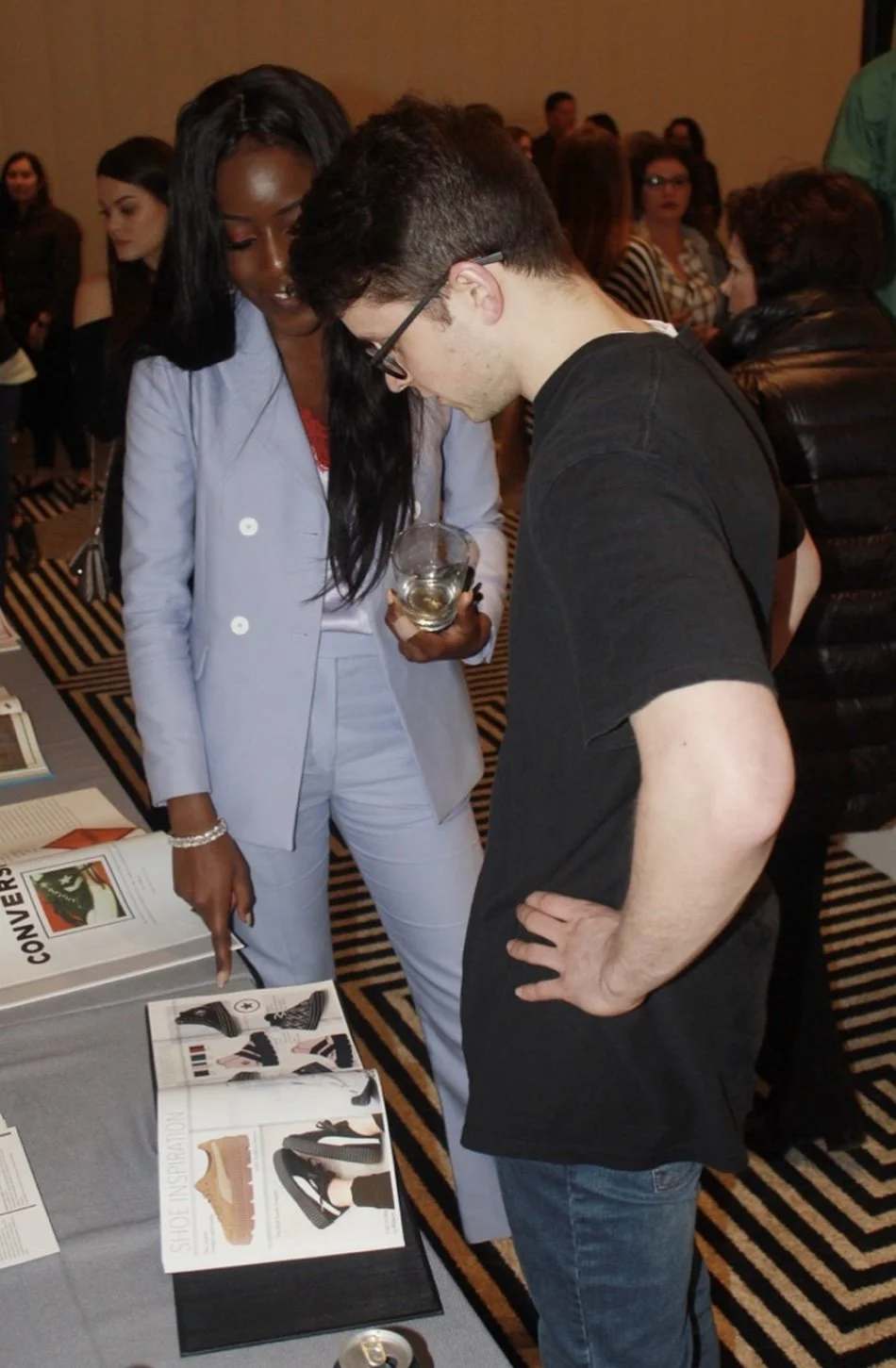 A woman in a light blue suit showing a shoe catalog to a man in a black t-shirt at an indoor event.
