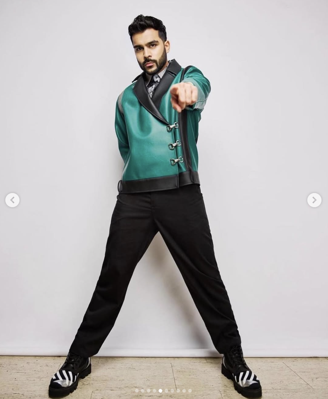 A man standing with legs apart, wearing a shiny green and black jacket, black pants, and black shoes with white stripes, pointing directly at the camera with a serious expression.