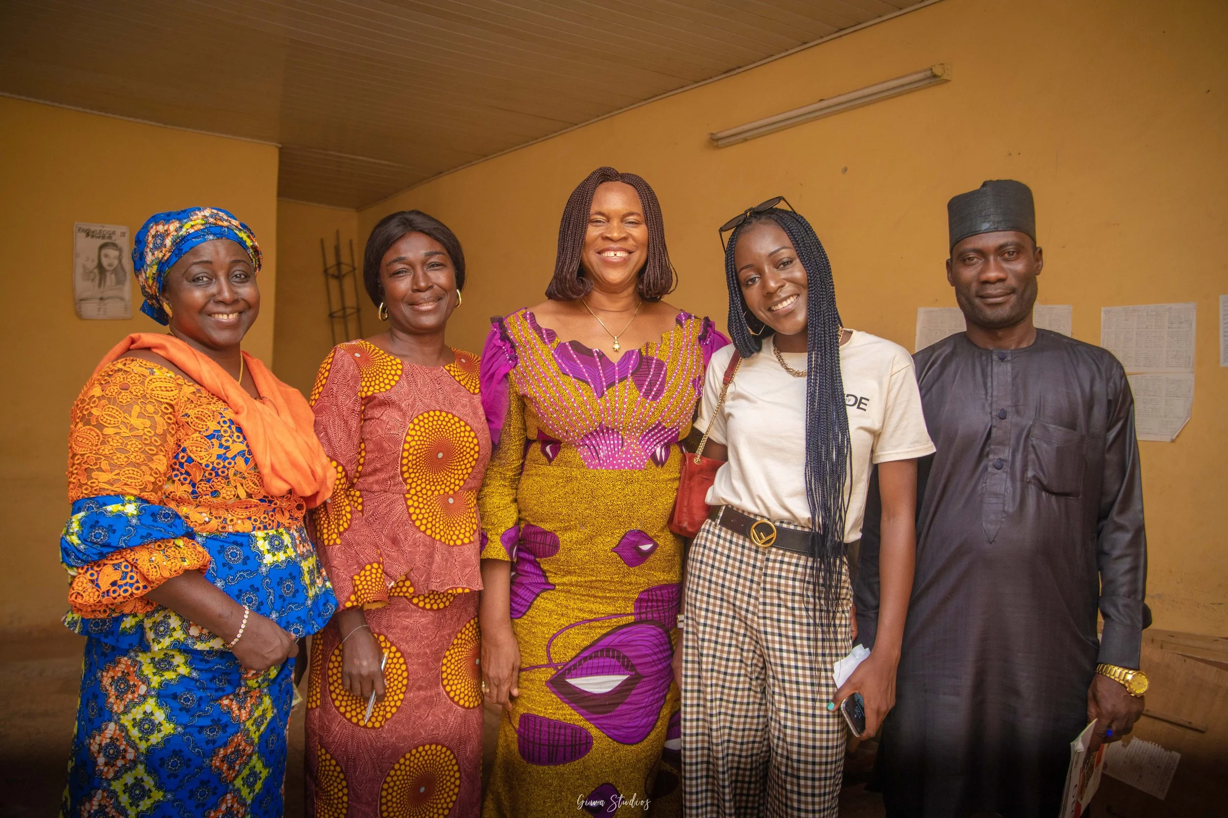 Group of six people standing together indoors, dressed in colorful and traditional African attire, smiling at the camera.