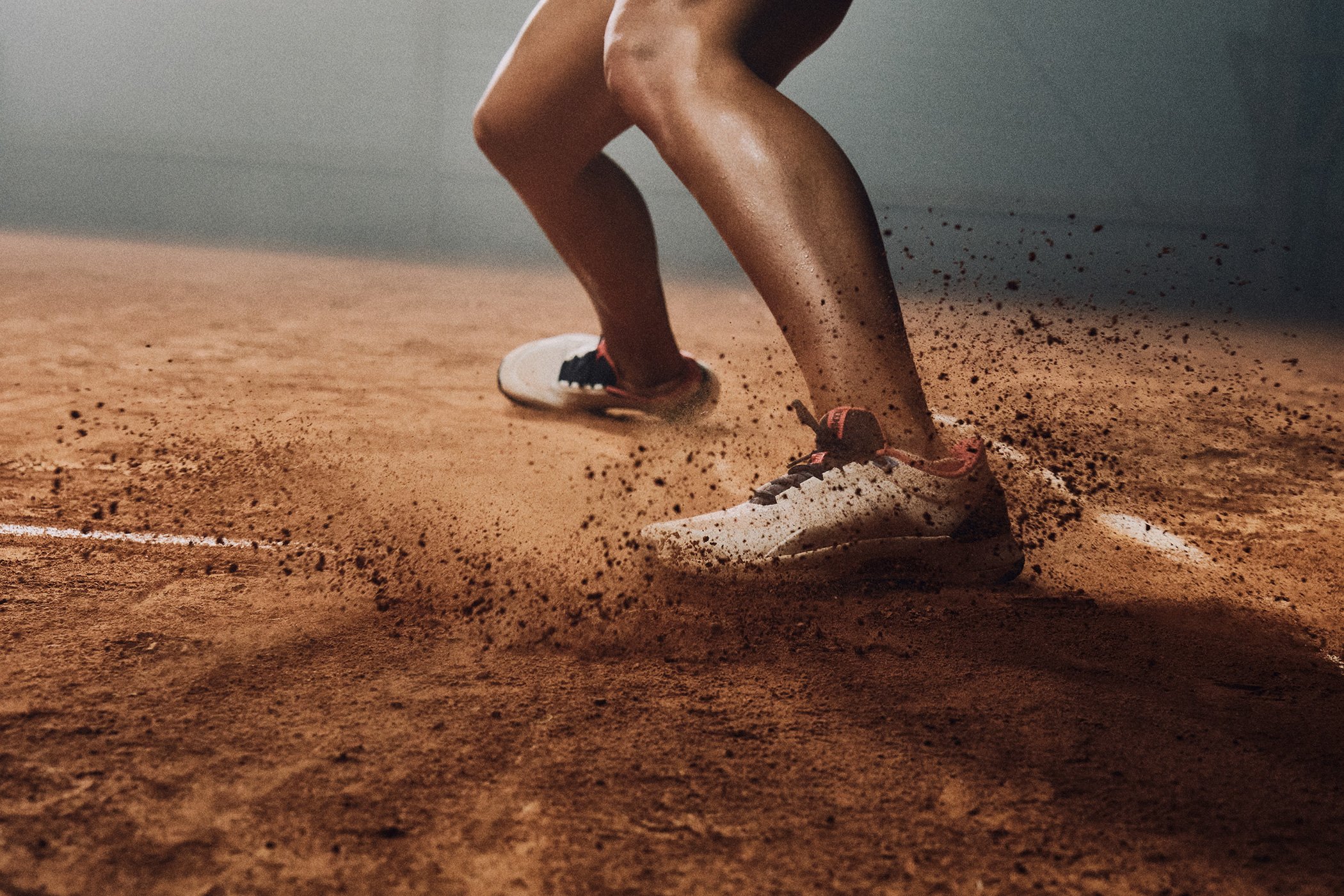 A person playing tennis on a clay court, wearing white tennis shoes and shorts, with a splash of dirt kicked up as they move.