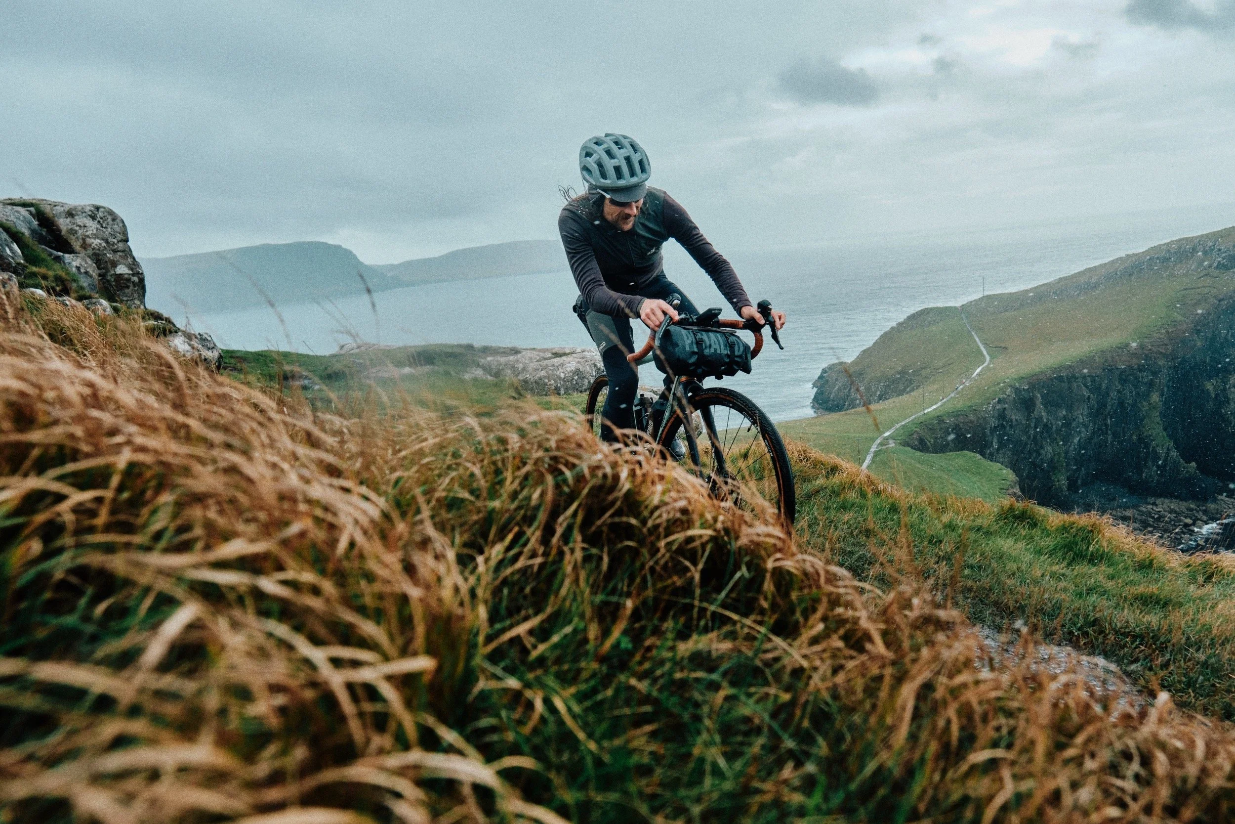 A person mountain biking on a grassy trail along a coastline with cliffs and water in the background on a cloudy day.