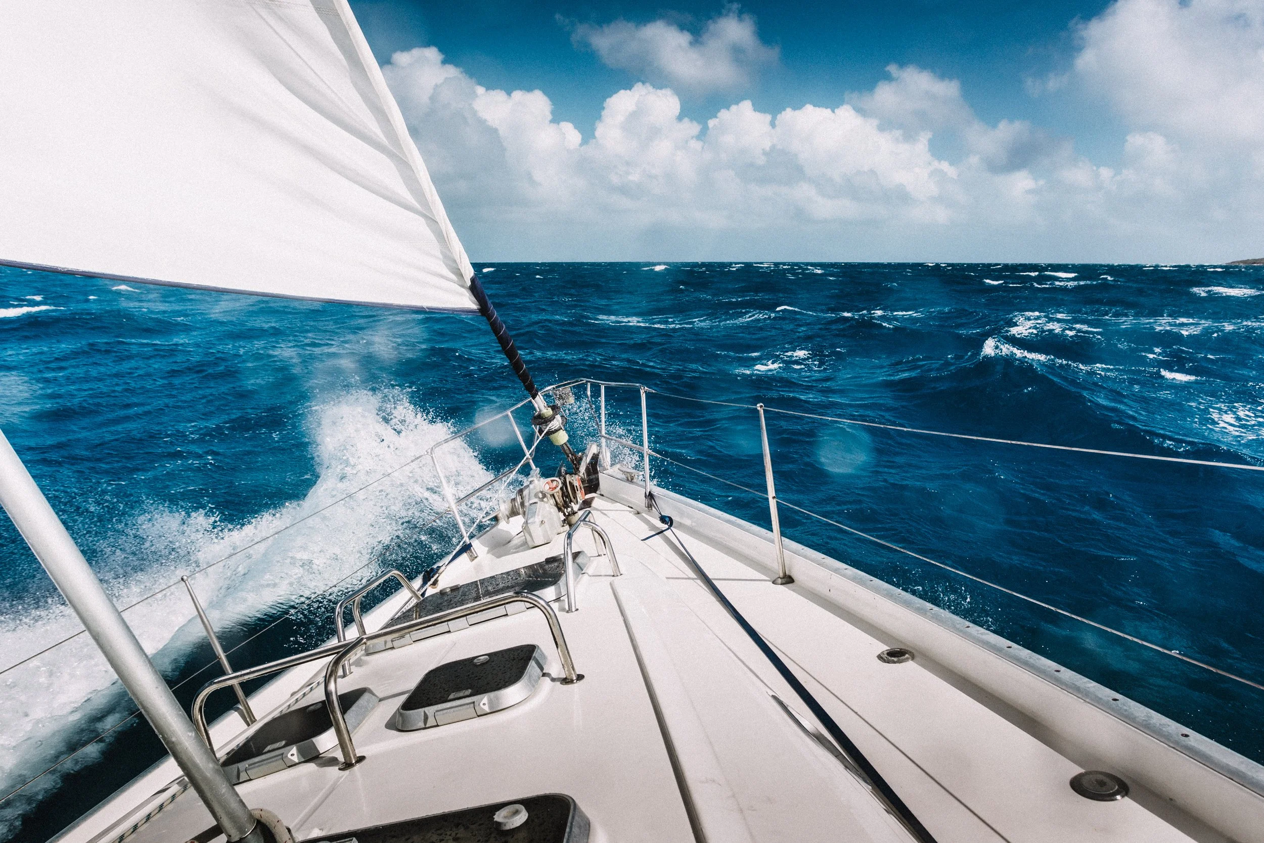 View from the bow of a sailboat on blue ocean water with white clouds in the sky.