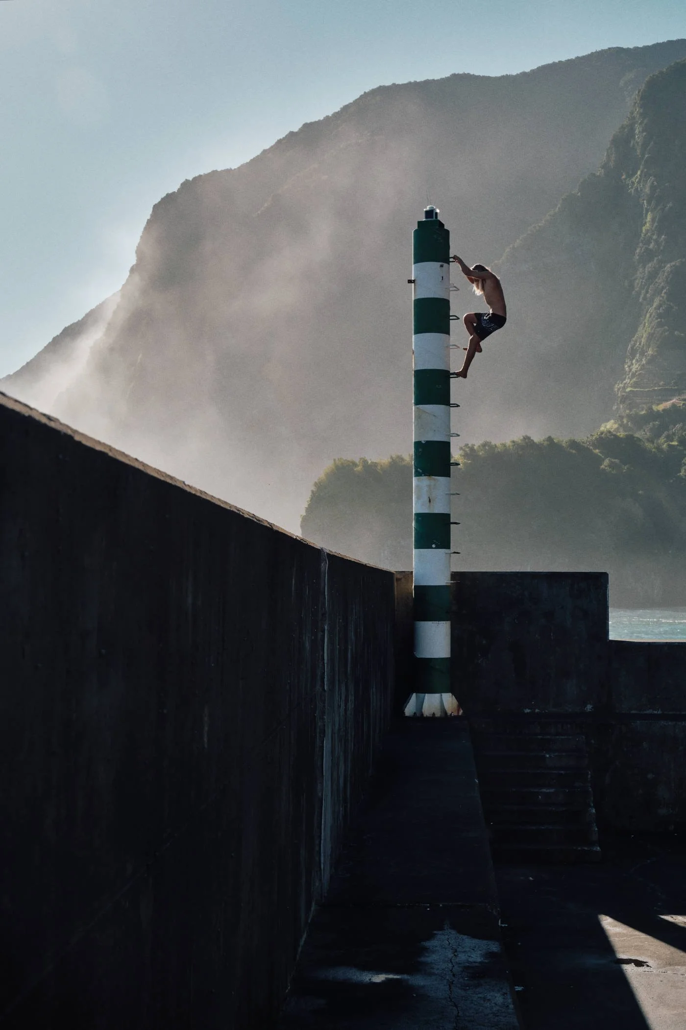 A person climbing a striped black and white lighthouse near a waterfront with mountains in the background.