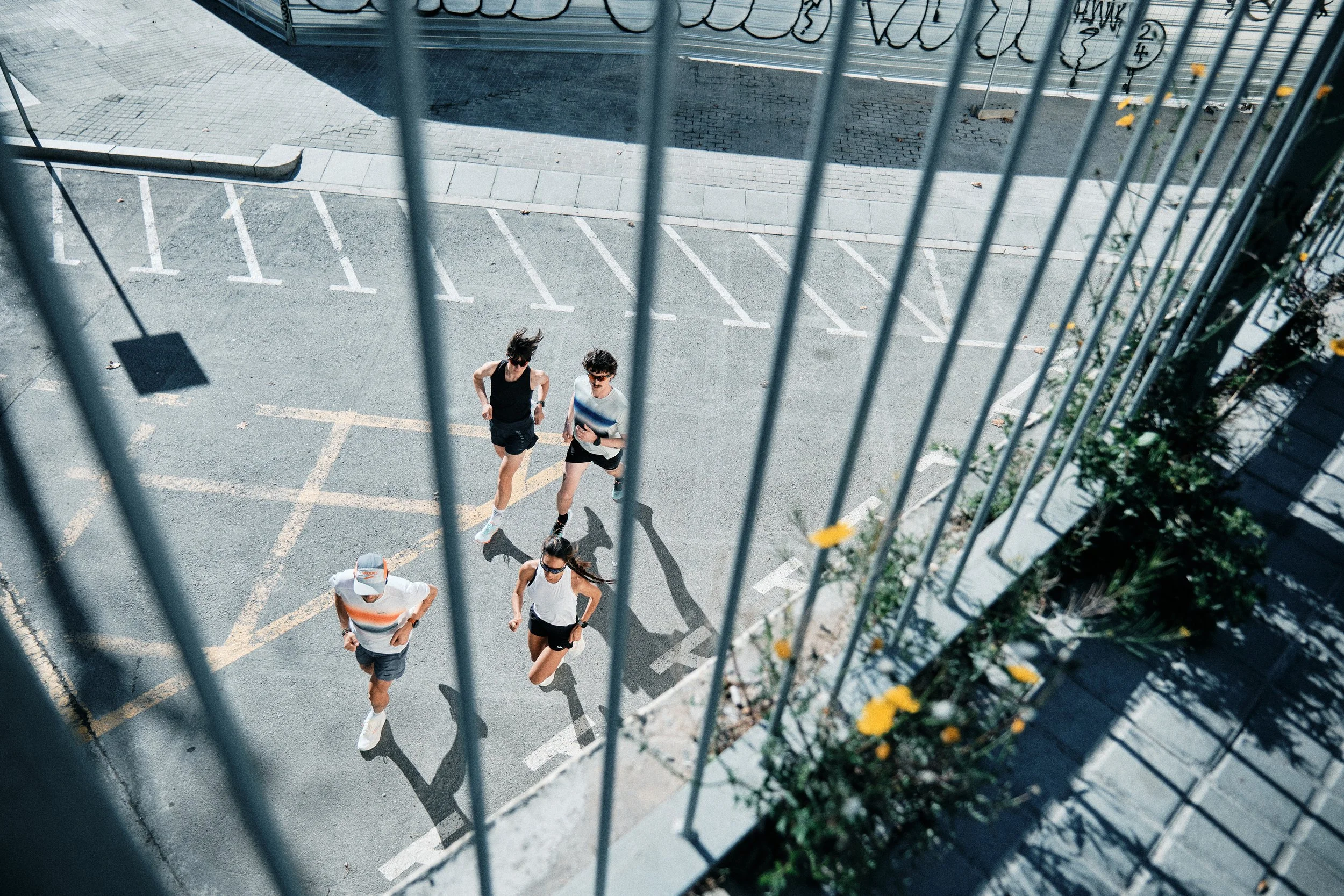 Four runners in athletic attire, two women and two men, jogging through a parking lot seen from an elevated perspective through metal bars of a fence.