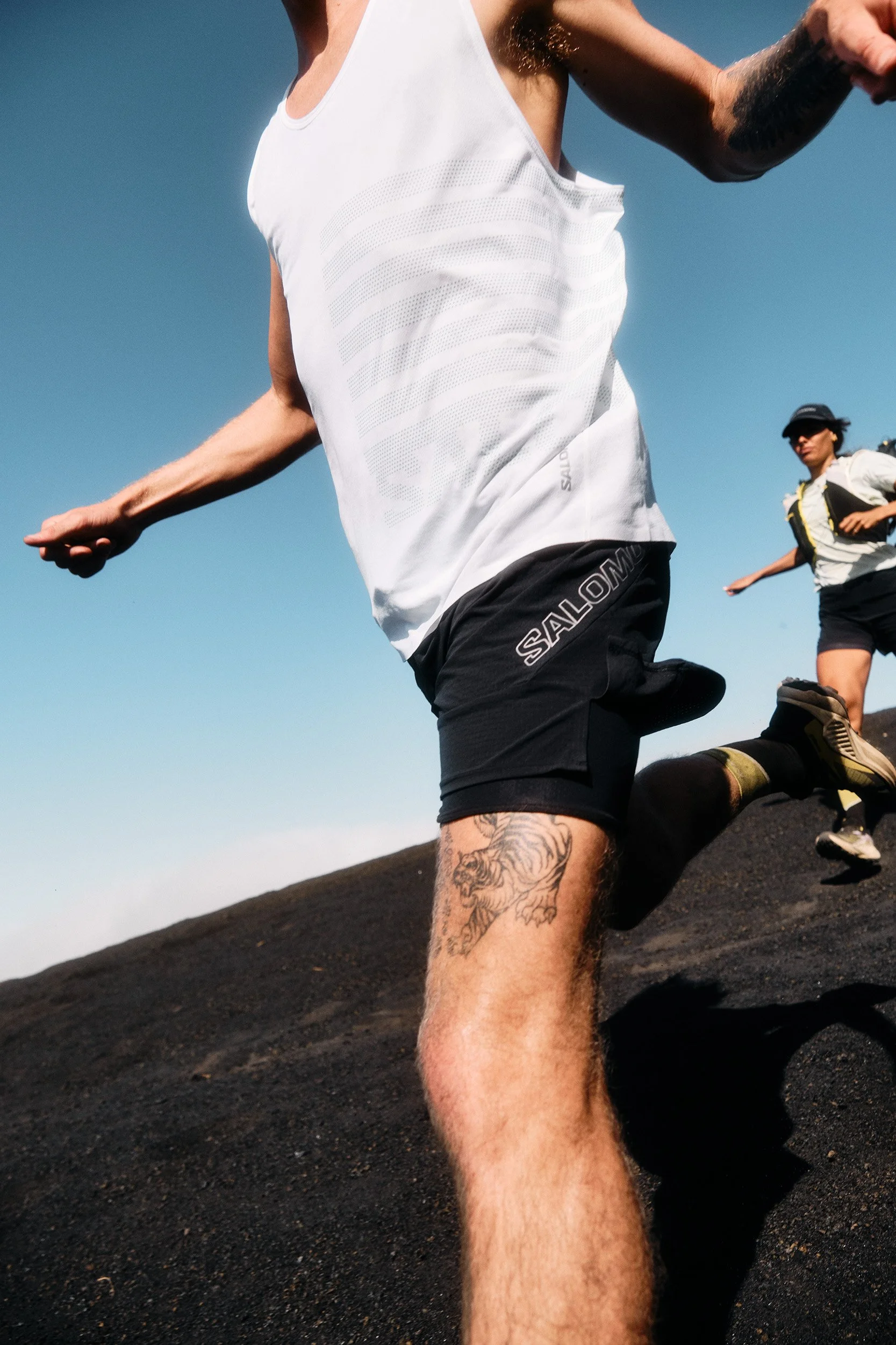 Close-up of a male trail runner in a white tank top and black shorts with a tattoo on his thigh, running uphill on a dark gravel trail under a clear blue sky, with a female trail runner in the background.