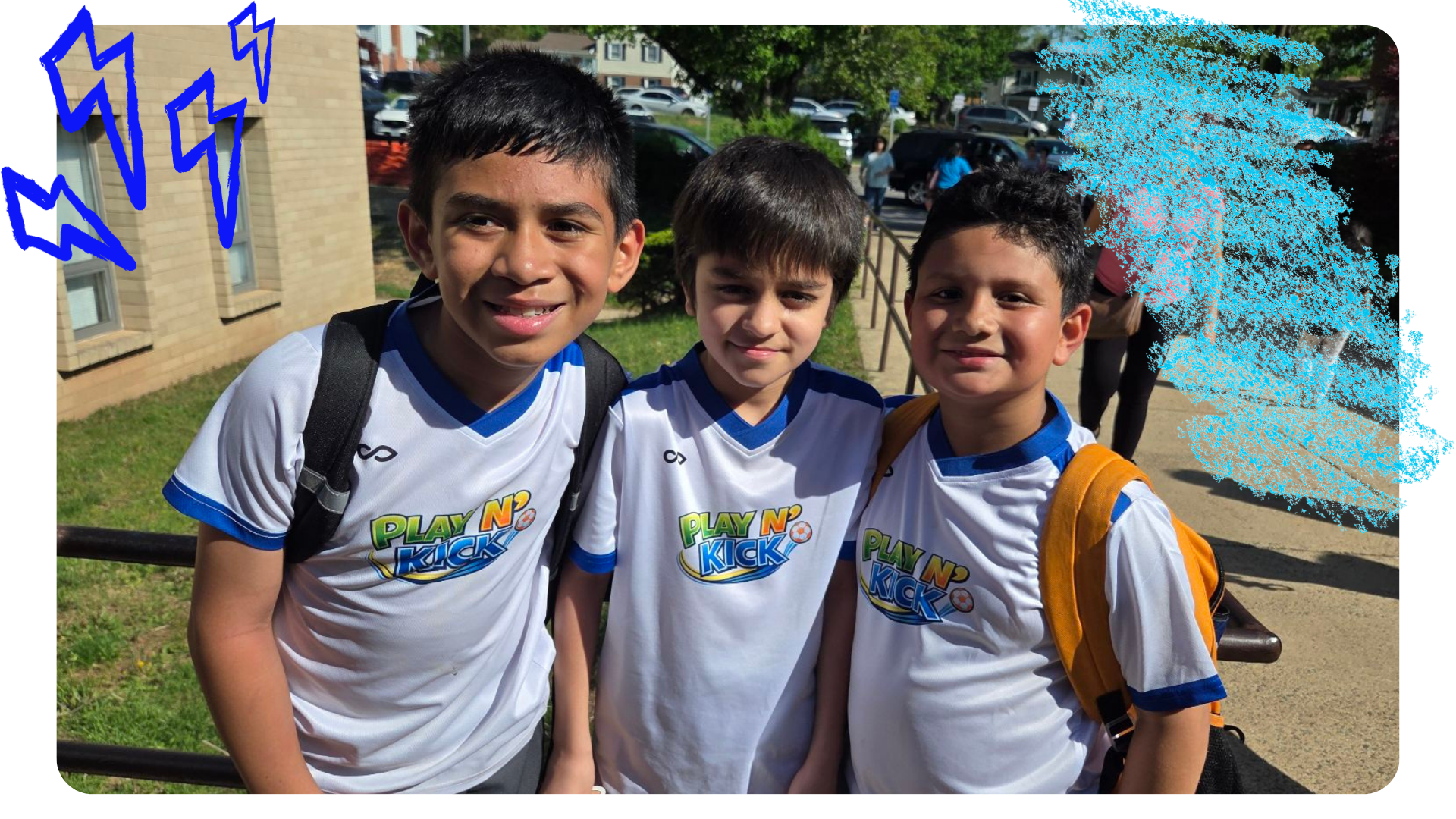 Three young boys wearing white and blue sports jerseys with 'PLAY N' KICK' logo standing outside on a sunny day, smiling for the camera.