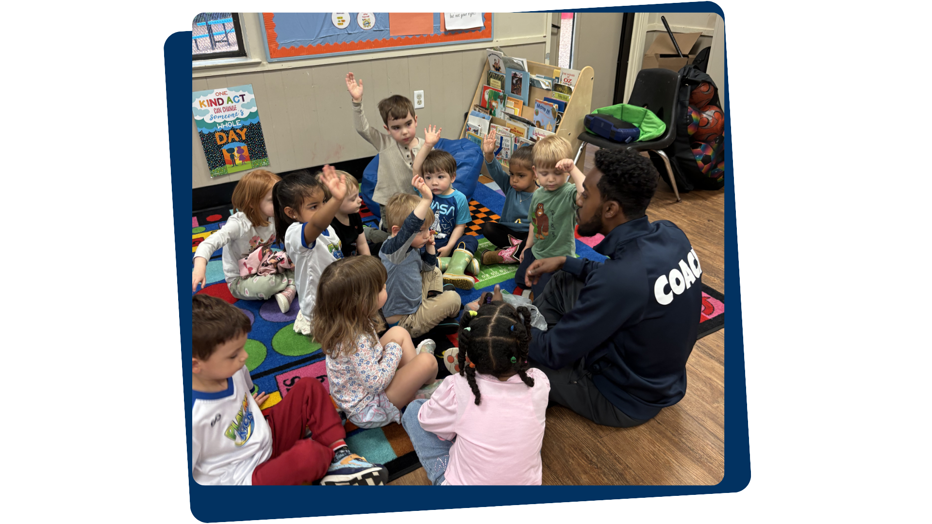 A teacher or coach sitting on the floor with a group of young children in a classroom setting. Some children are raising their hands. There are bookshelves, a colorful rug, and educational posters in the background.