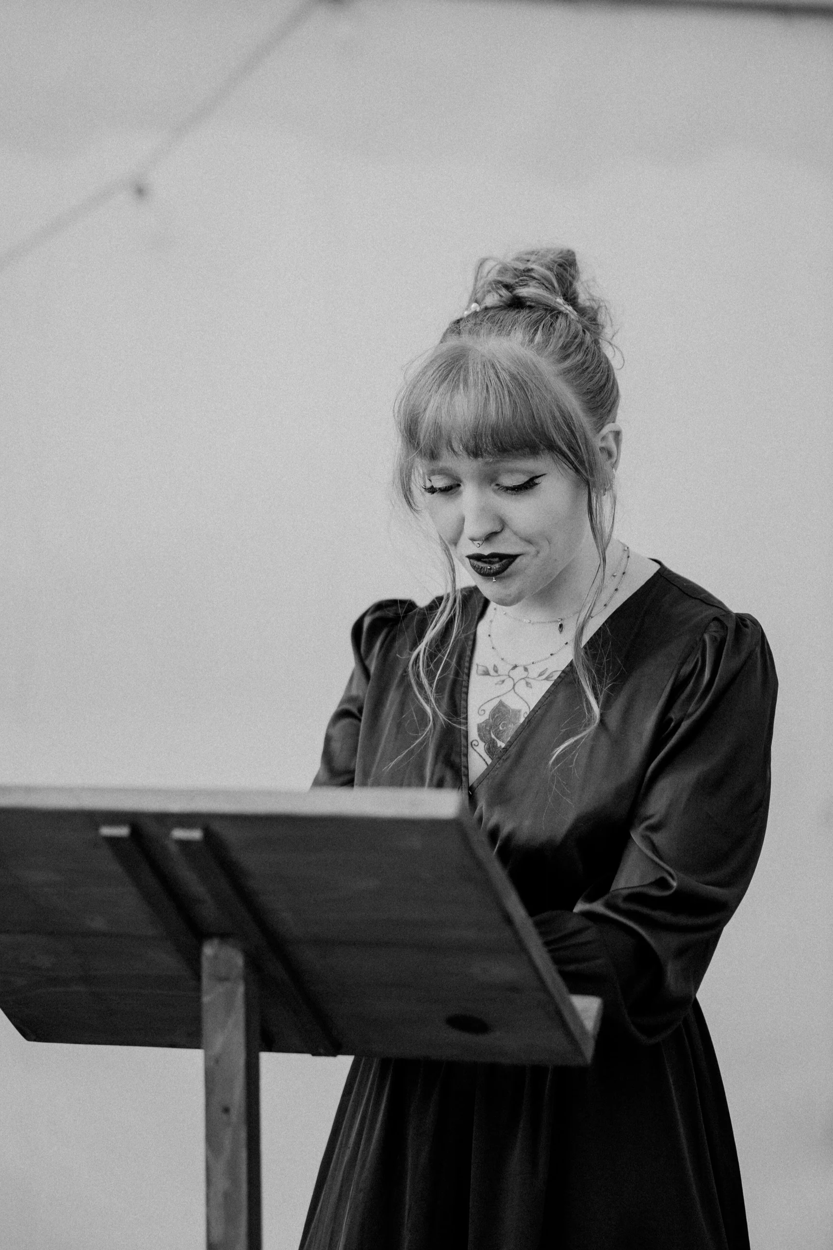 Une femme avec des tatouages, des cheveux en chignon et un maquillage distinctif regarde vers le bas, debout devant un pupitre en bois.