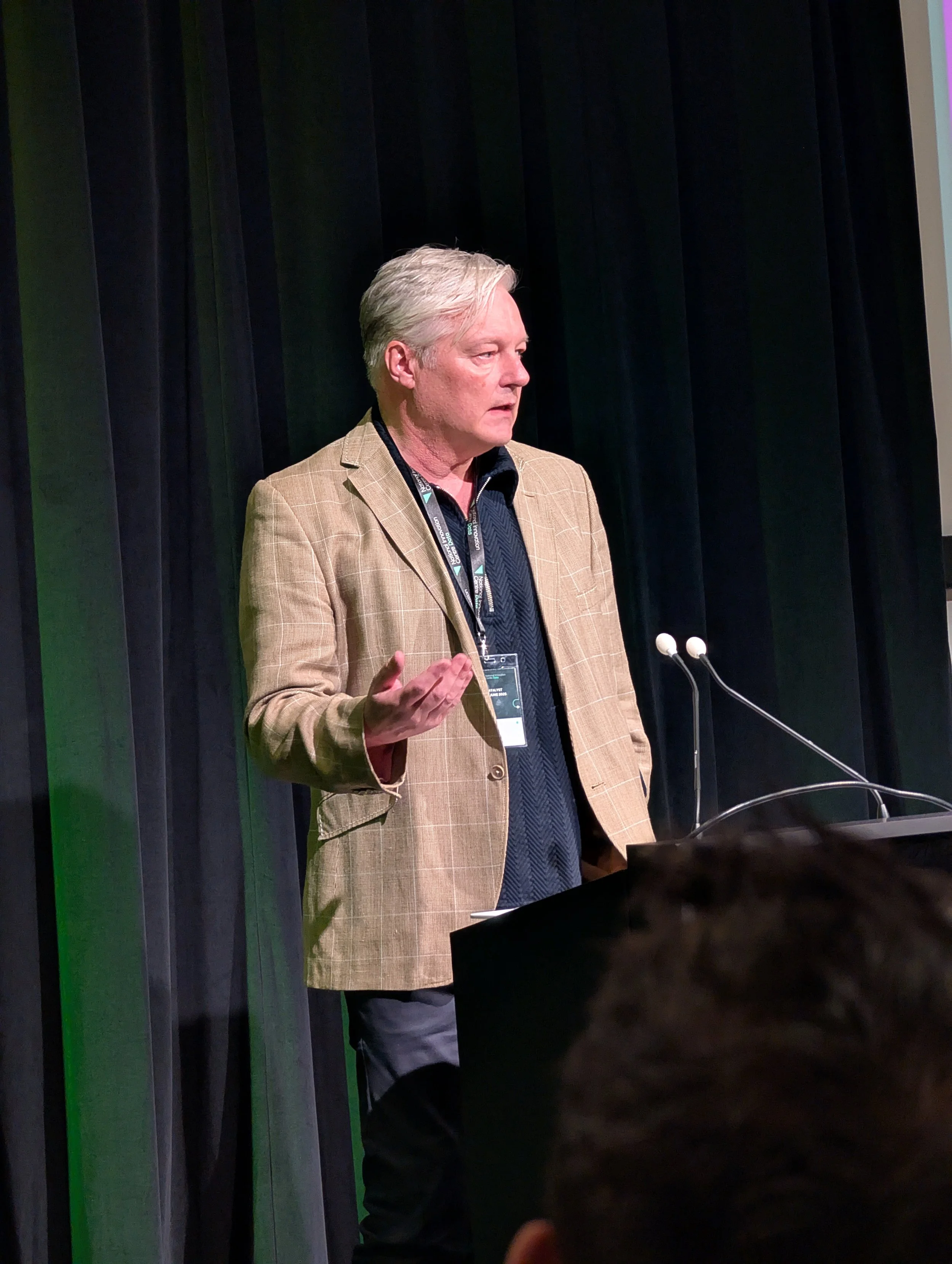 A man in a tan checkered blazer giving a presentation or speech at a podium with microphones, black curtains in the background, and a person in the audience in the foreground.