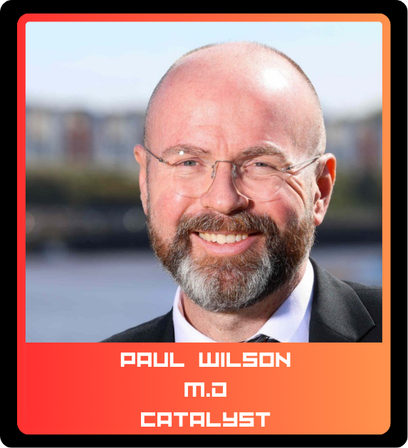 Close-up photo of Paul Wilson, a man with a beard and glasses, smiling outdoors with a blurred cityscape and water in the background.