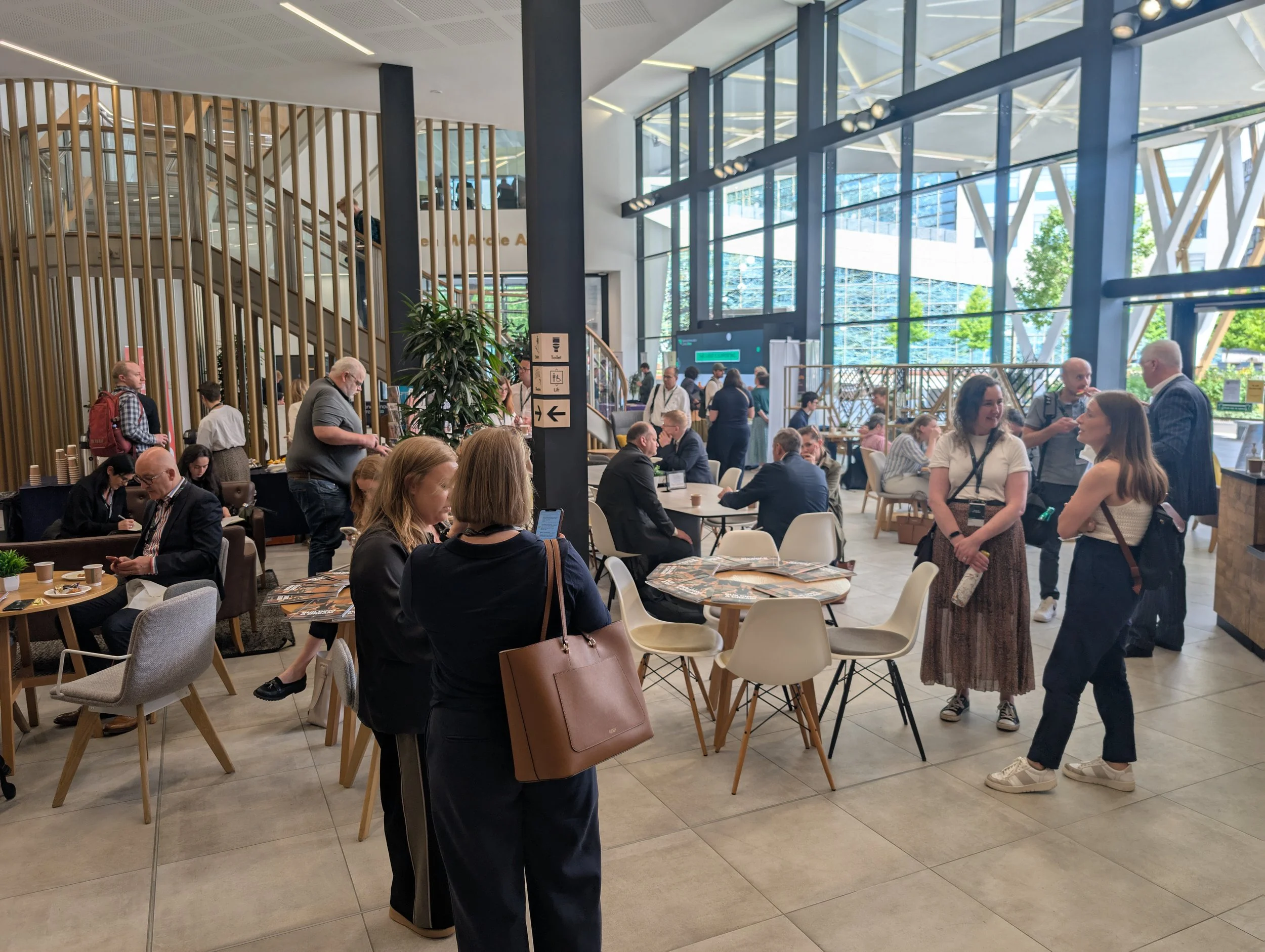 Crowd of people in a modern, spacious indoor area with large glass windows, wooden accents, and seating, likely at a conference or event venue.