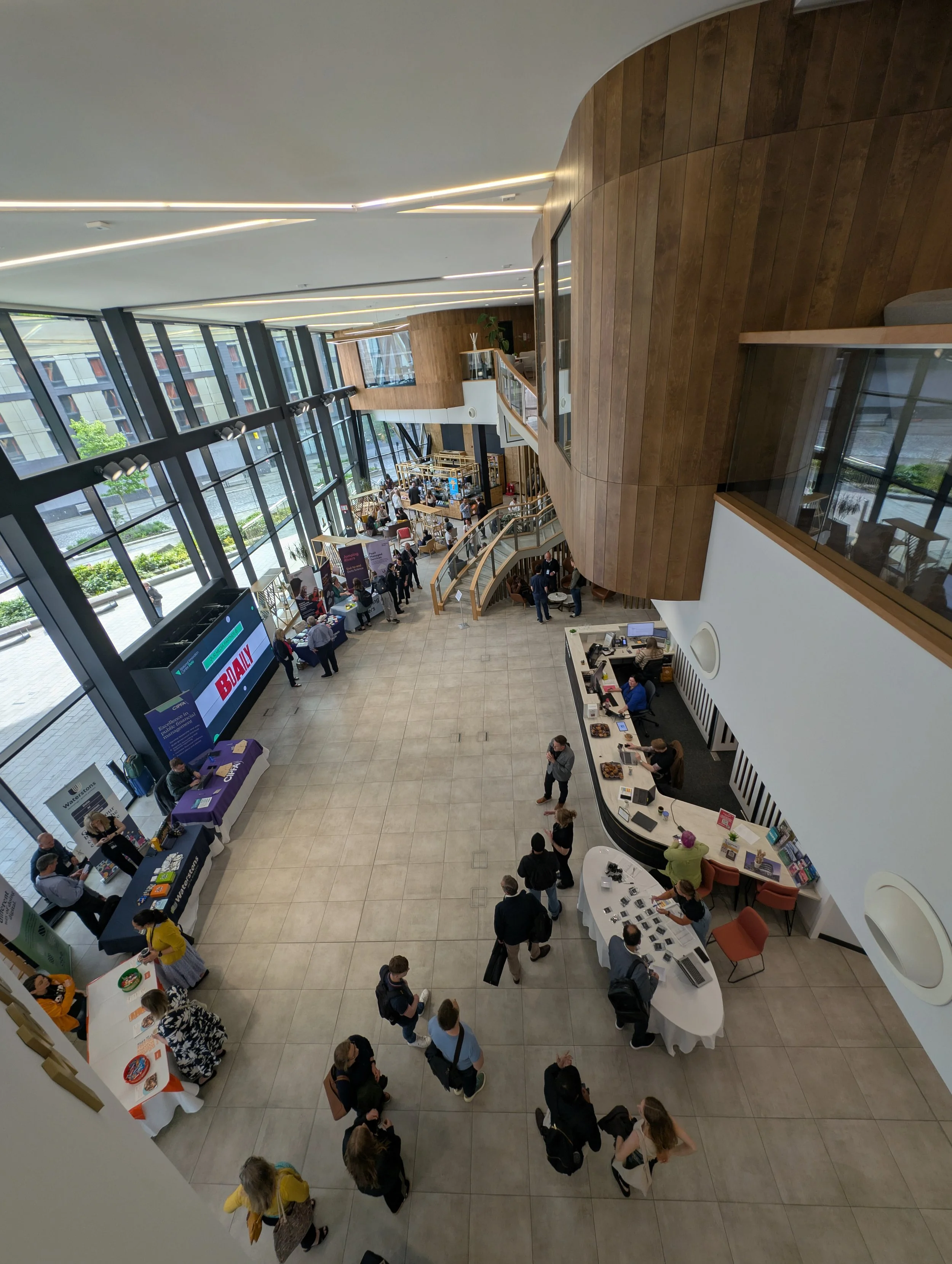 Interior view of a modern, spacious lobby with high glass windows, a reception desk, seating area, and several people attending an event or conference.