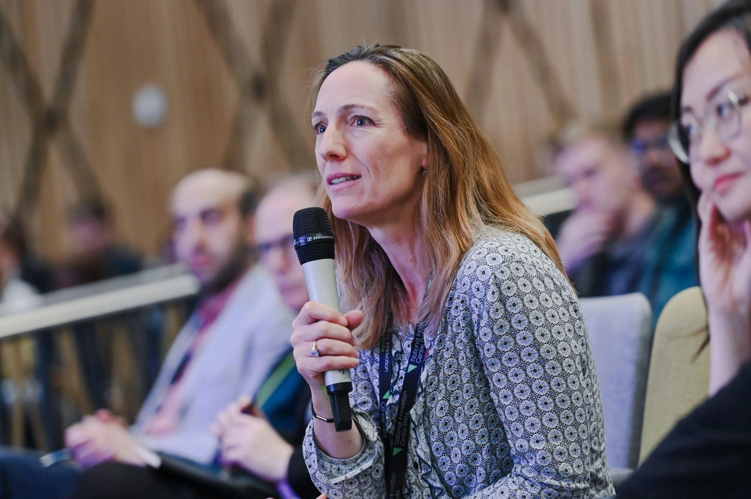 A woman with light brown hair holding a microphone during a conference or panel discussion, with other people sitting behind her in an audience.