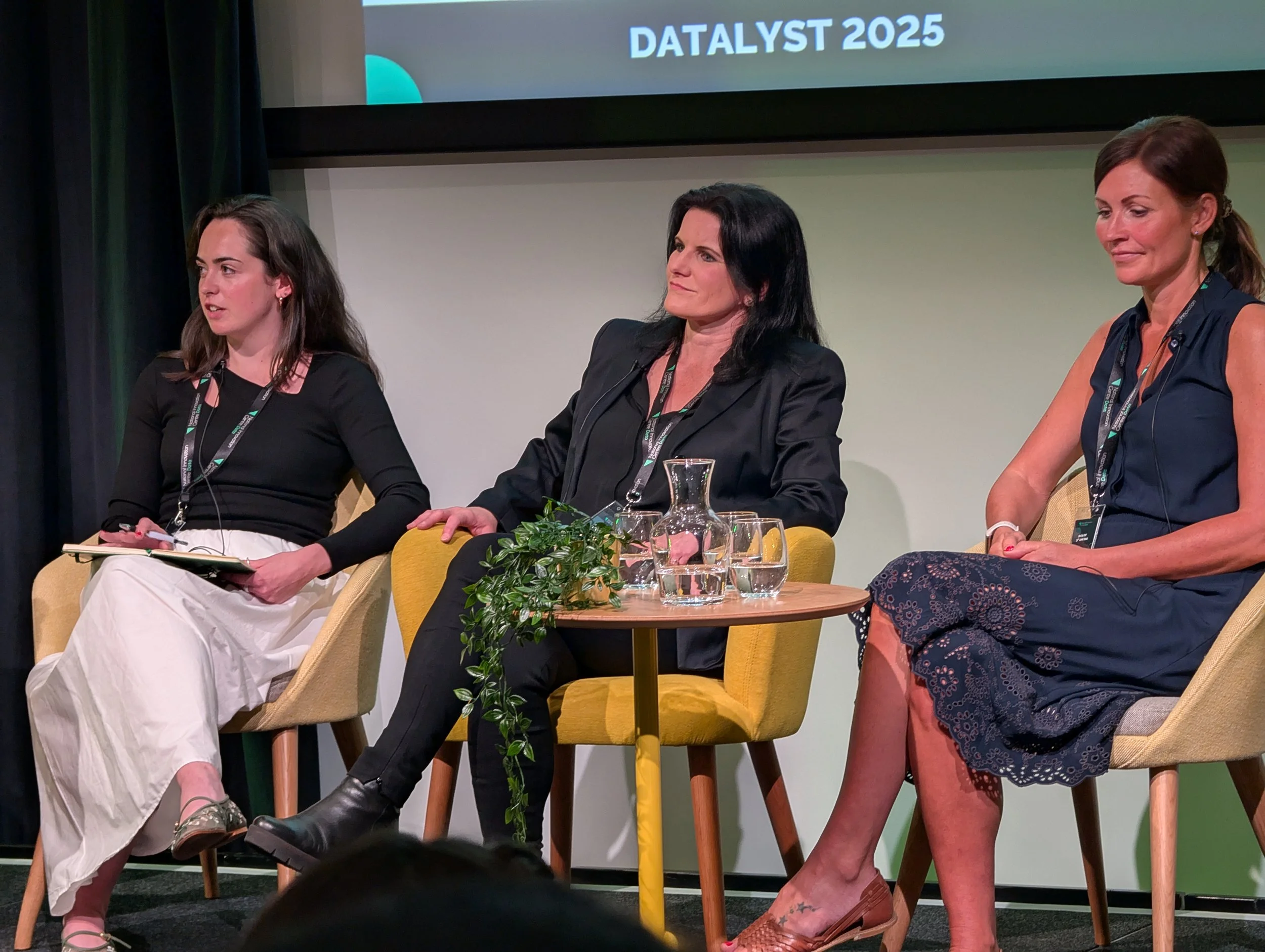 Three women sitting on stage at a panel discussion during DATALYST 2025 event, with a small table holding water glasses and a plant in front of them.