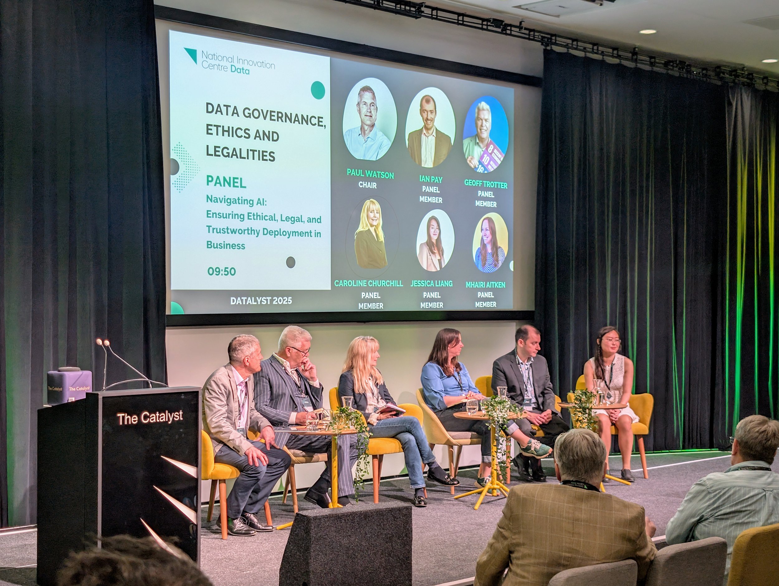 A panel discussion at a conference with six speakers sitting on stage, and a large screen behind them displaying panel information, including speaker photos and titles. The screen shows the panel topic: 'Data Governance, Ethics and Legalities,' and f