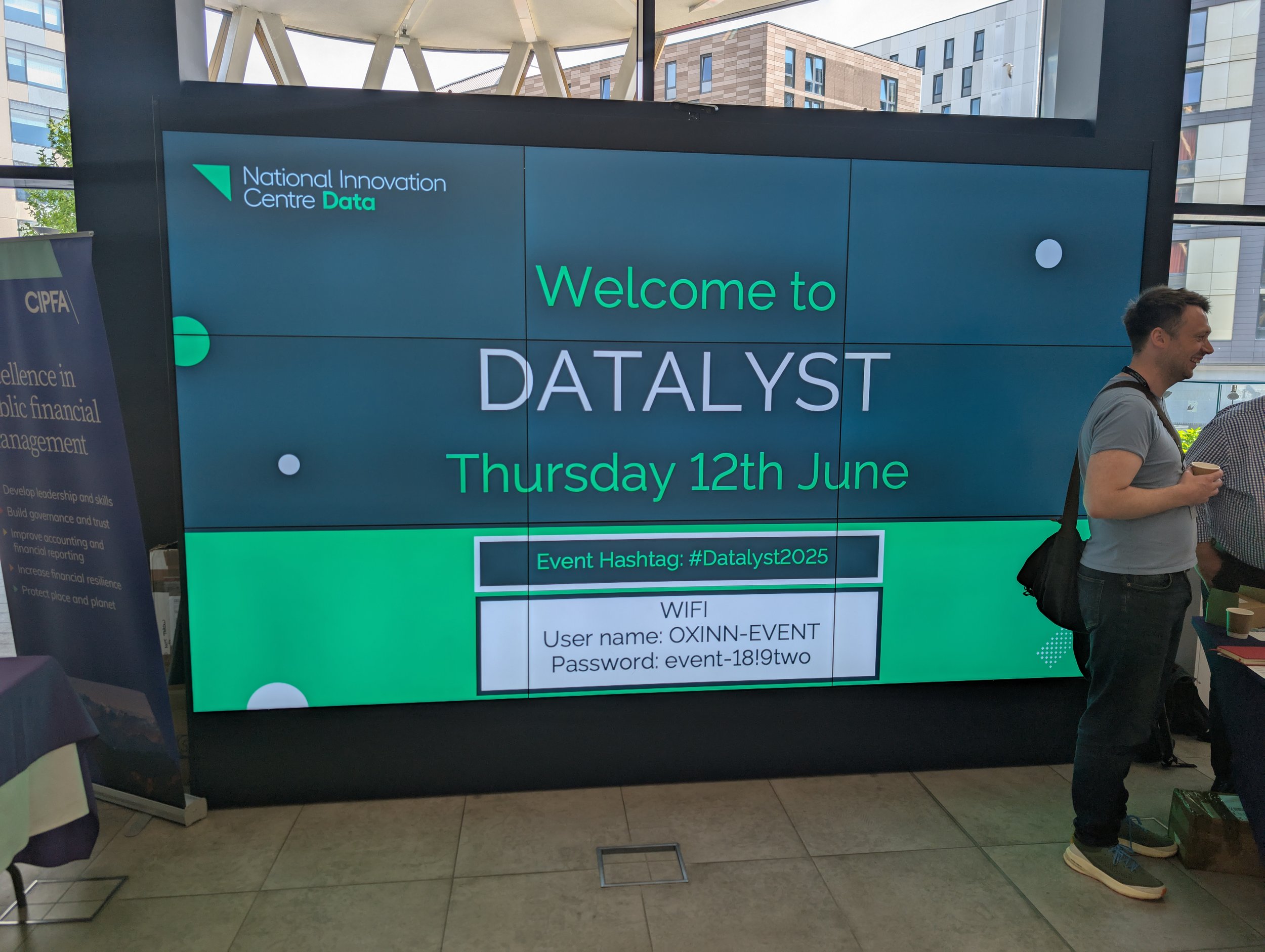 Large digital screen displaying an event welcome message for DATALYST on Thursday, June 12th, 2023, with Wi-Fi details and event hashtag #Datalyst2025, situated in a modern building with large windows and people standing nearby.