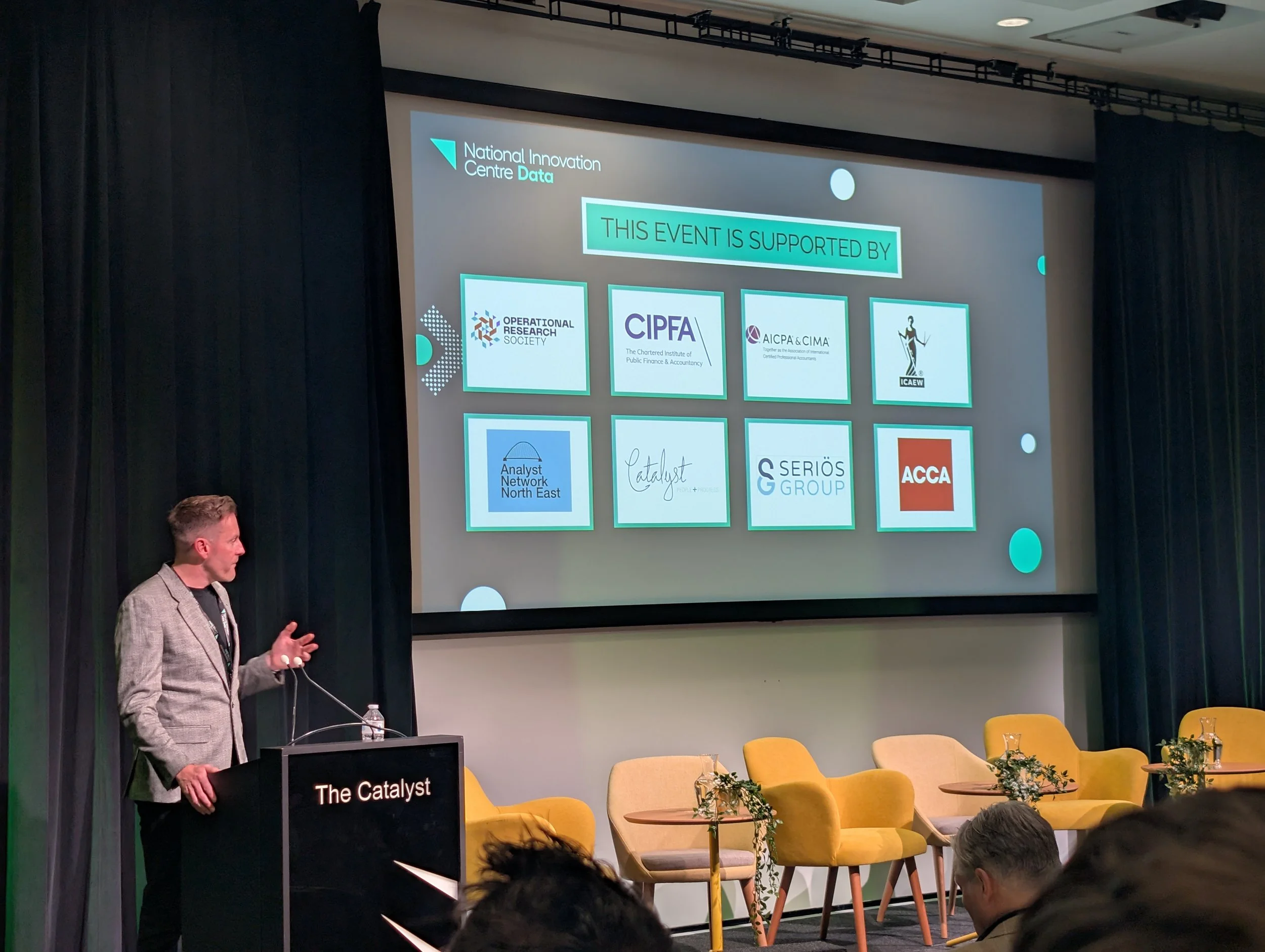 Man in a gray blazer giving a presentation at a conference with a slide showing logos of supporting organizations, including Operational Research Society, CIPFA, ICAEW, and others, on a large screen behind him.