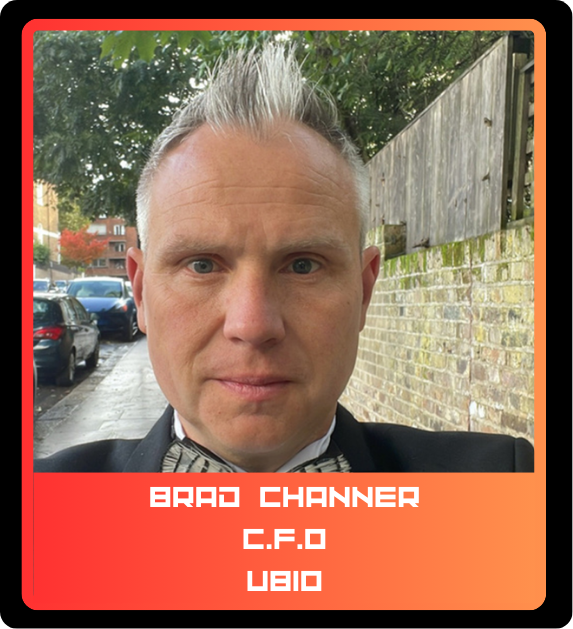 Close-up of a man with short gray hair, wearing a tuxedo and bow tie, standing outdoors on a city street with parked cars and brick walls in the background.