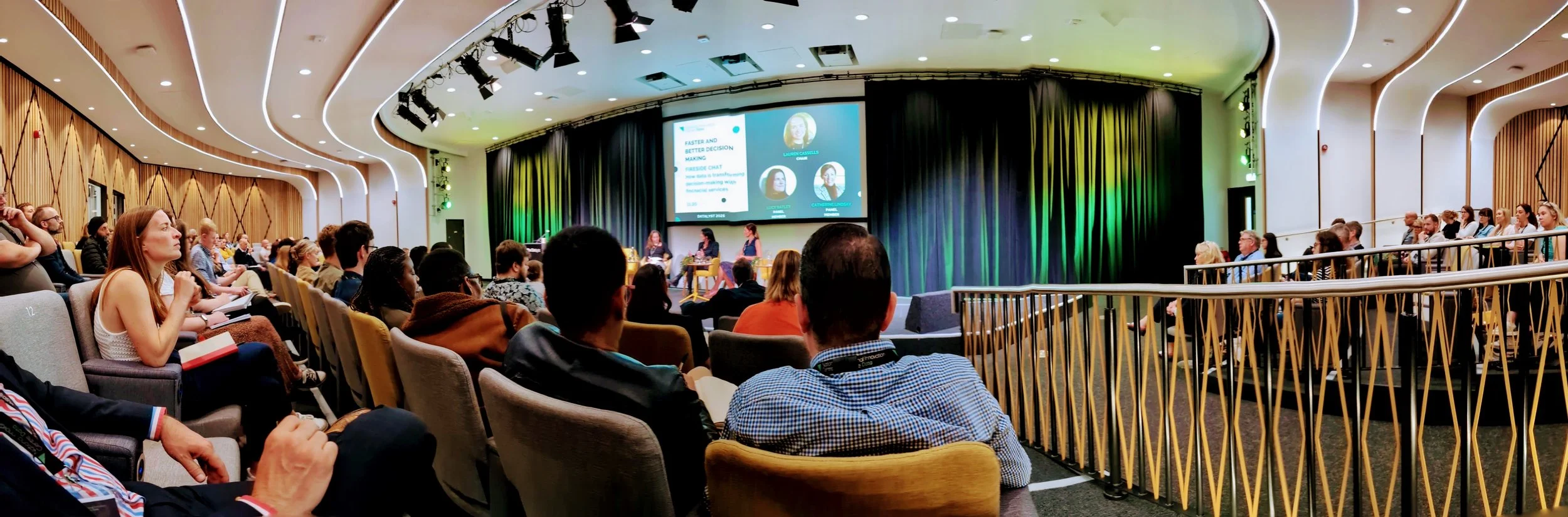 Audience seated in a modern auditorium watching a panel discussion on stage with a large screen displaying presentation slides.