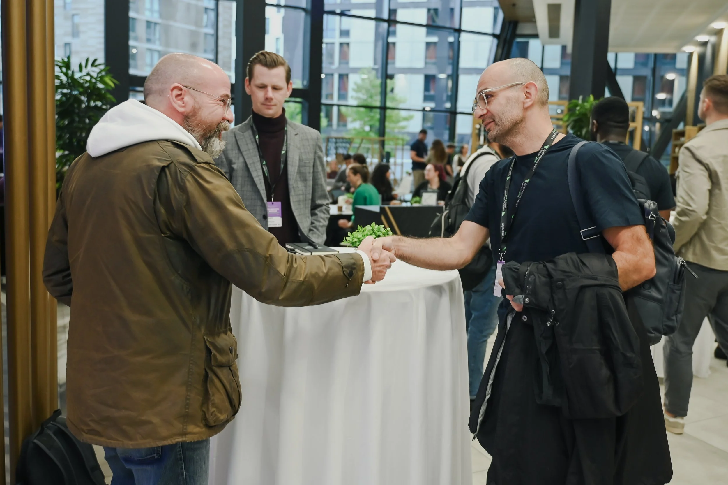 Two men shaking hands at a conference or networking event, with several other attendees in the background inside a modern building with large glass windows.