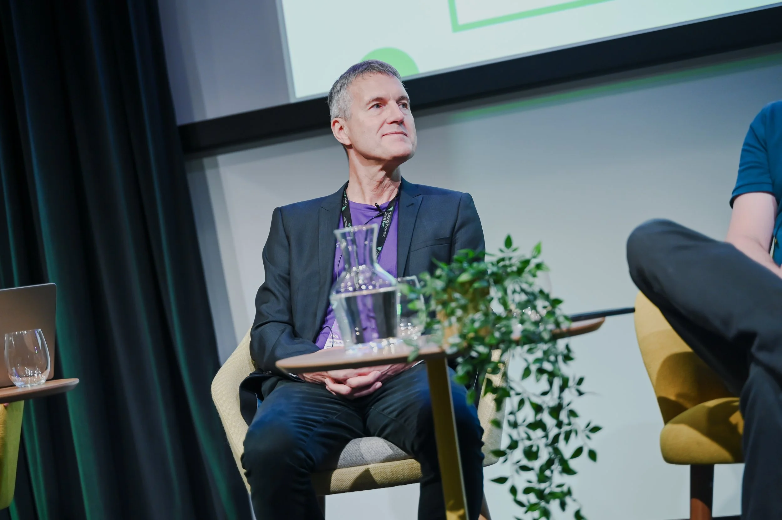 A man sitting on a beige chair during a panel discussion, with a table in front of him holding a glass pitcher, a glass, and some greenery. He is wearing a dark blazer and purple shirt.