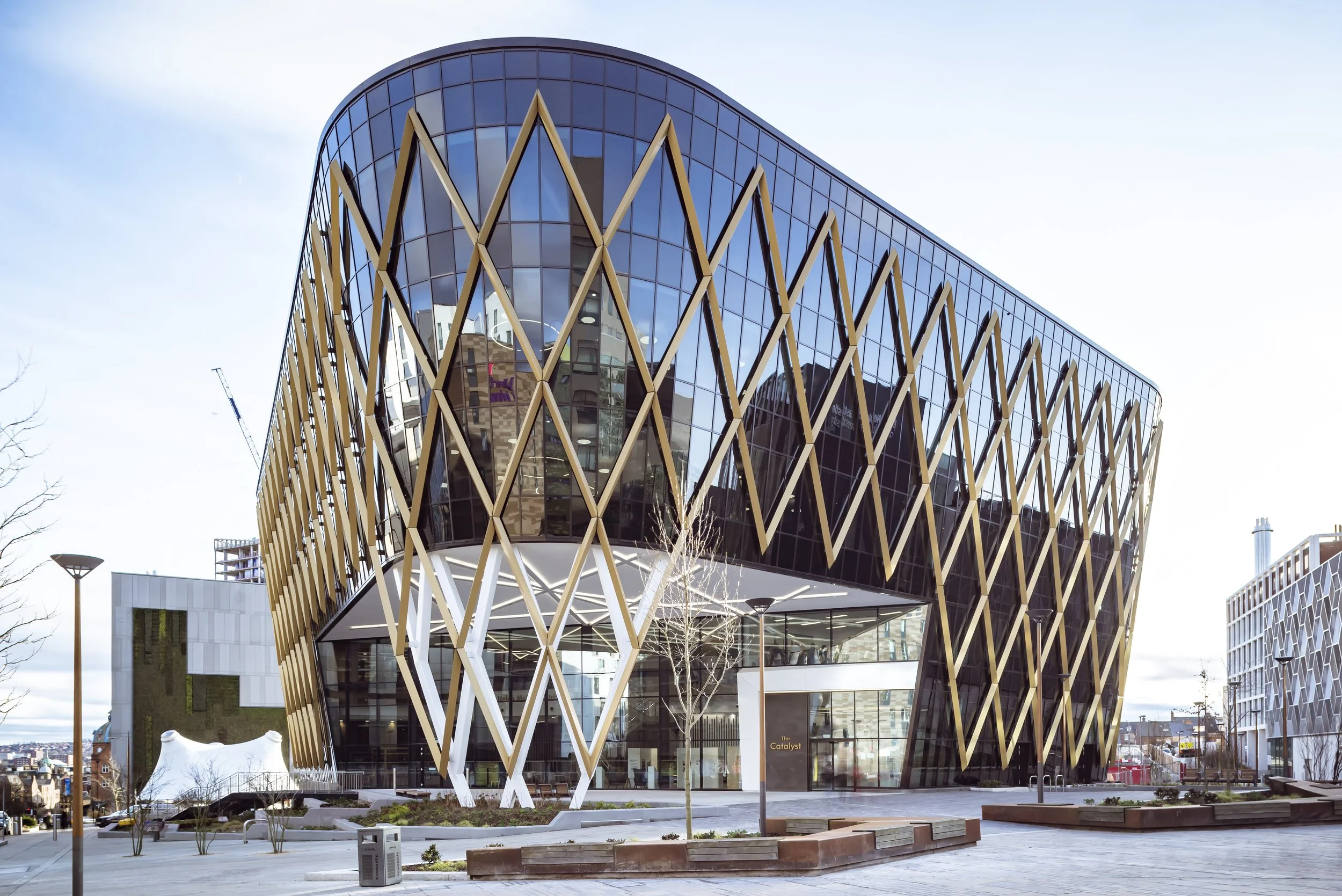 Modern building with a glass exterior and gold lattice design, situated in an urban area with benches and trees in the foreground.