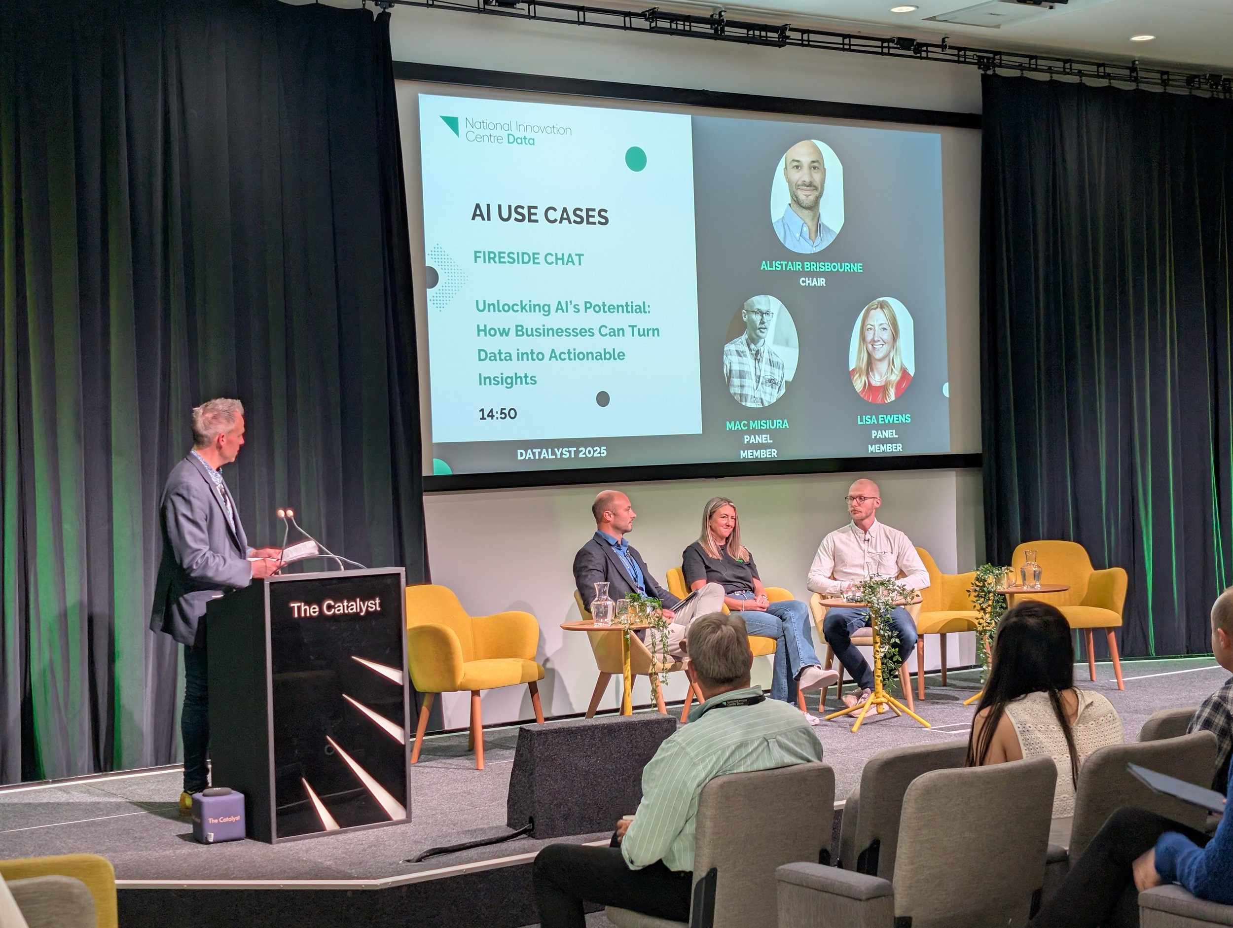 A panel discussion at a conference with four participants, three seated on stage and one standing at a podium. A large screen behind the panel displays a presentation about AI use cases, featuring headshots and names of the panelists, under a black c
