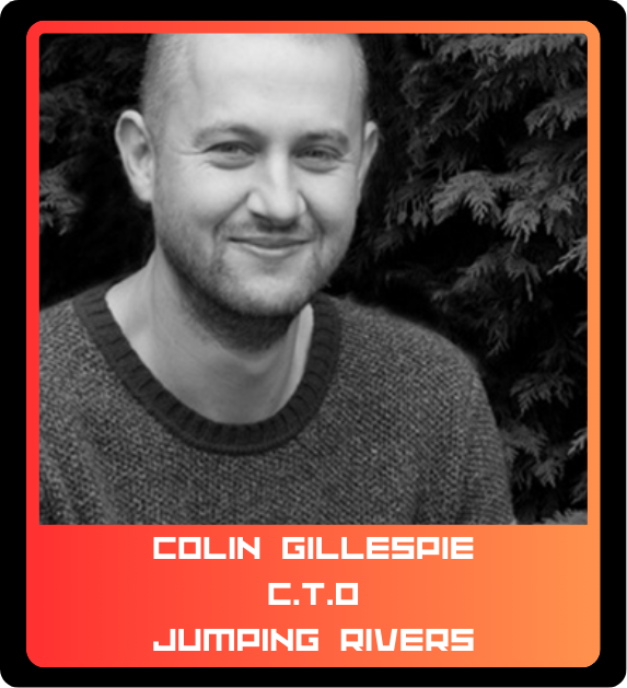 Black and white portrait of a smiling young man with short hair and a beard, wearing a sweater, standing in front of a bush or tree with foliage.