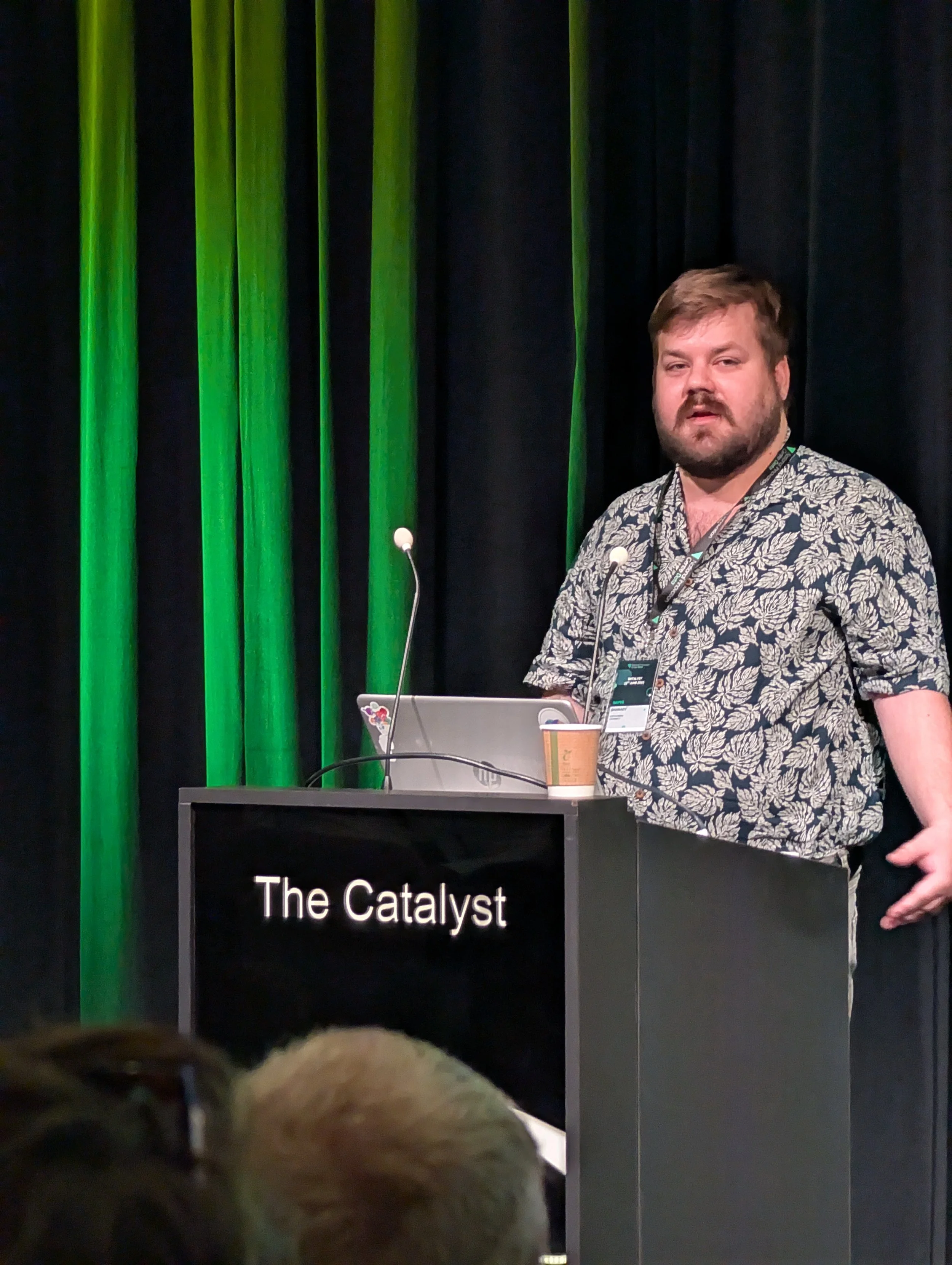 A man with a beard and wearing a patterned shirt standing behind a podium labeled 'The Catalyst,' with a laptop and coffee cup on top, in front of a black curtain with green stage lights.