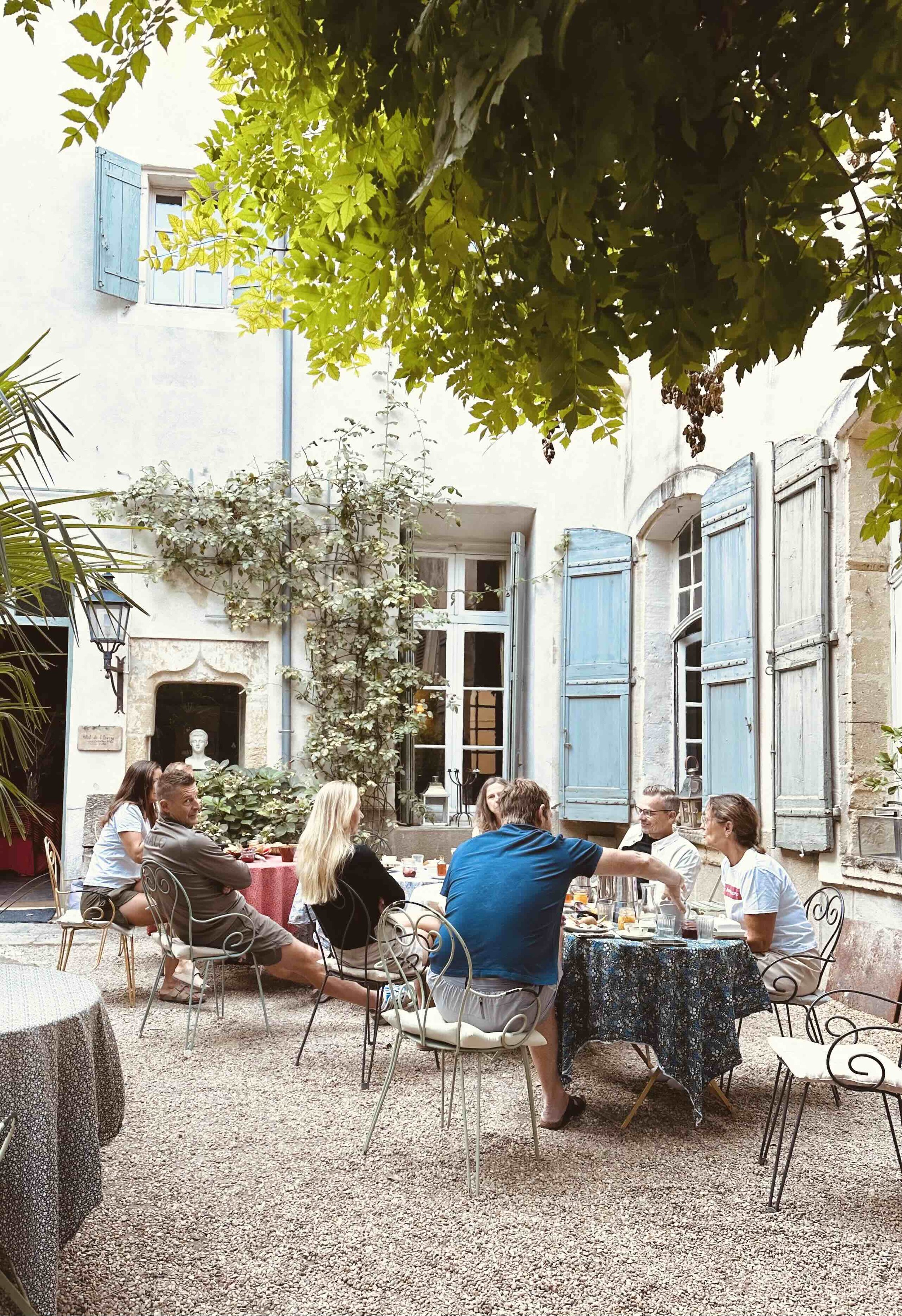 People having a meal outdoors in a courtyard with blue shuttered windows and greenery.