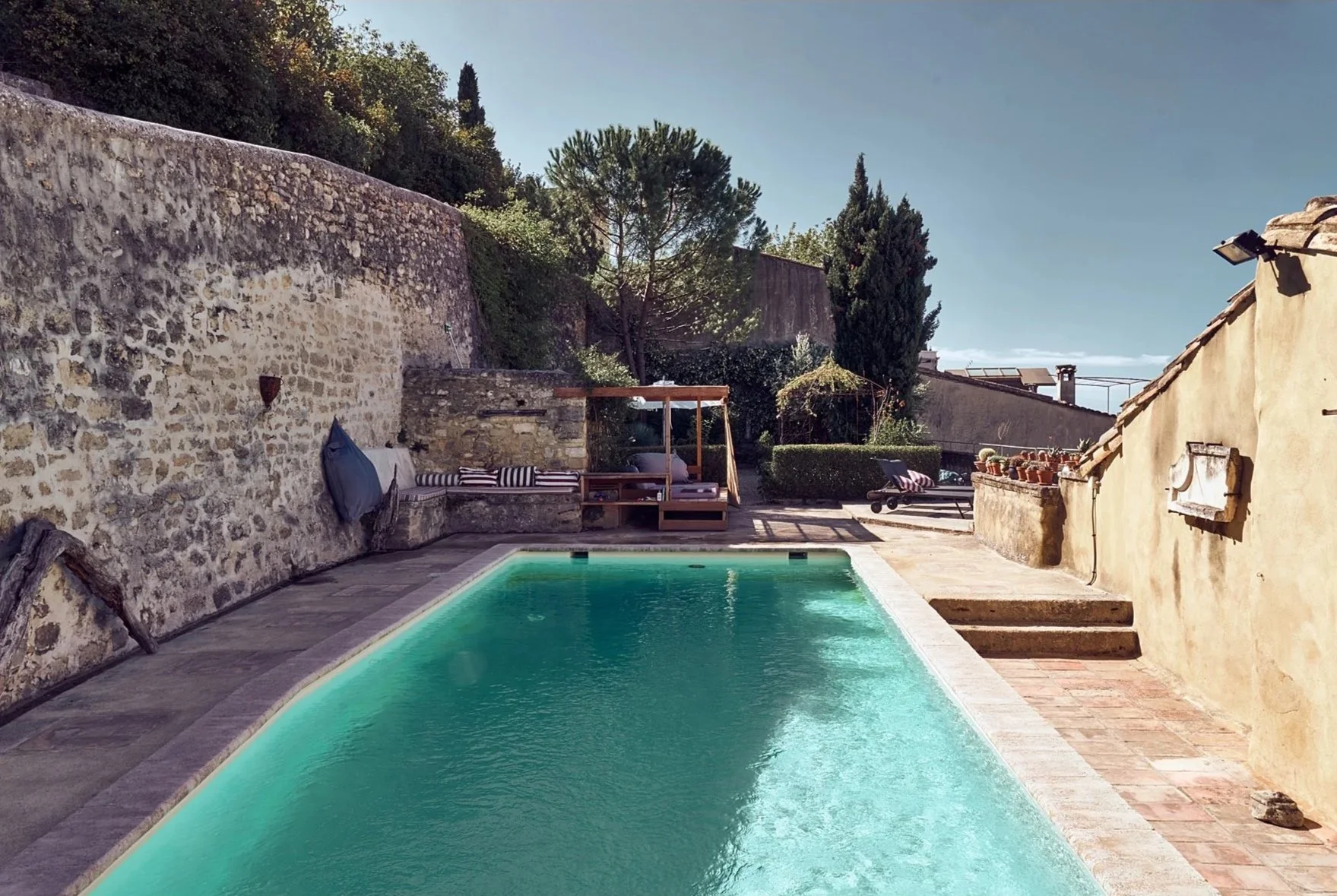 the perfect retreat a getaway spot in France with a swimming pool, stone and stucco walls, outdoor lounge area with striped cushions, trees, and a clear sky.