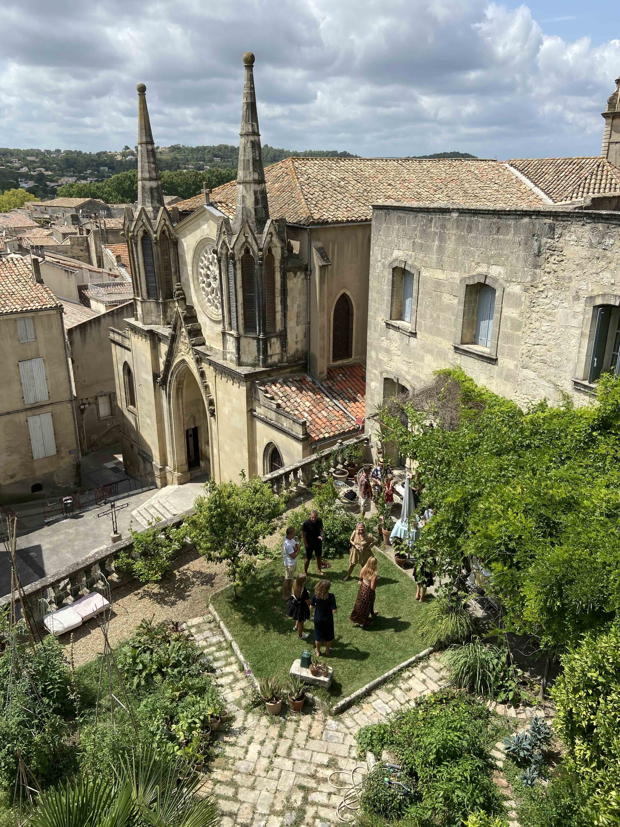 A church with two tall steeples and a stained glass window, located in an old European city with tiled roofs. In the courtyard, people are gathered, standing on grass and cobblestone pathways, surrounded by trees and potted plants, enjoying a social event on a partly cloudy day.
