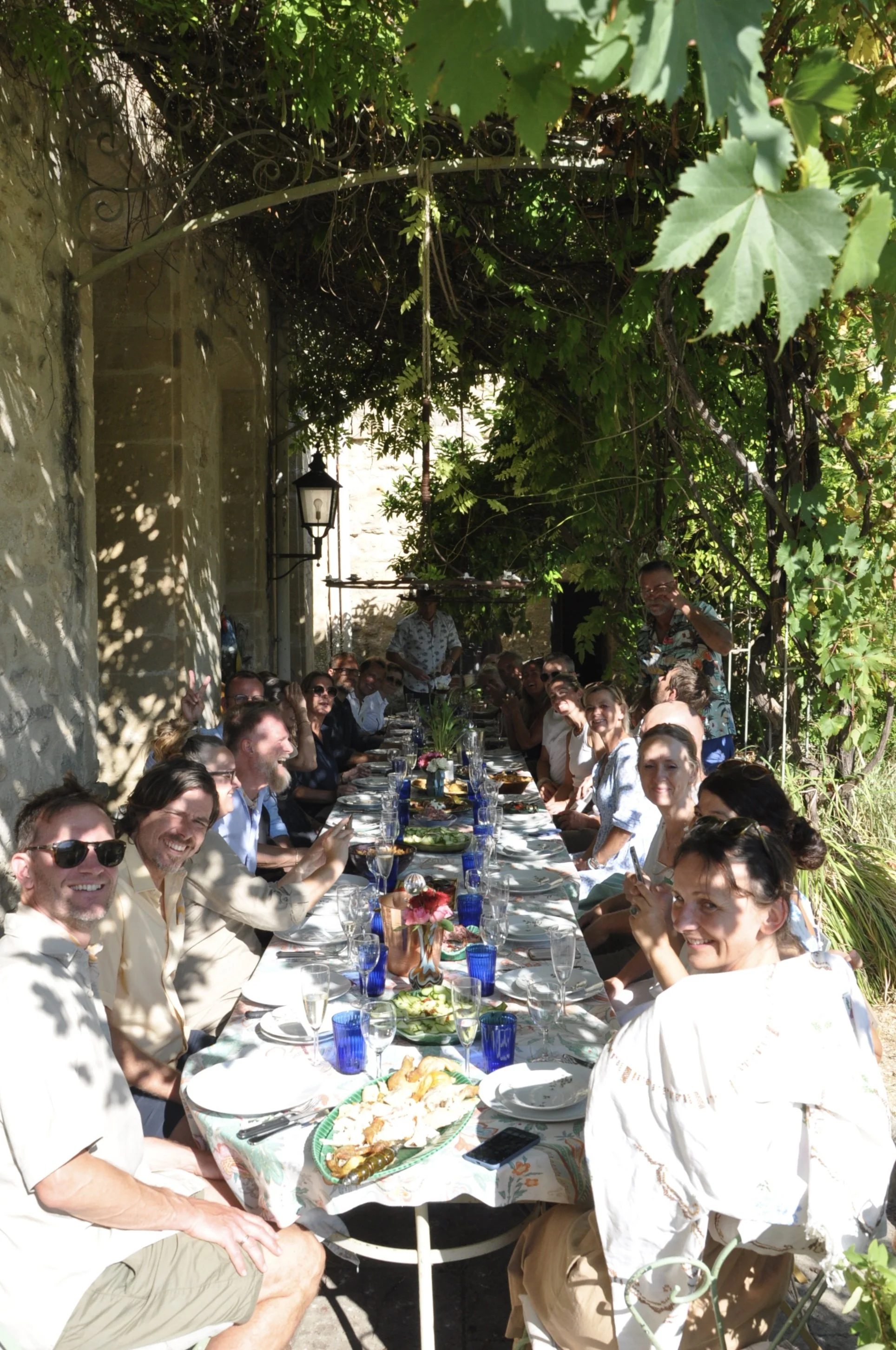 People gathered for outdoor brunch or lunch at a long table under a canopy of leaves, with food and drinks on the table.